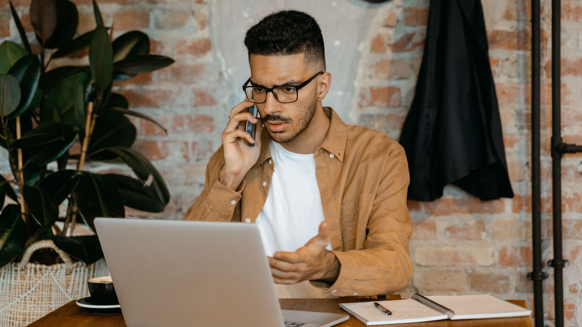 A man in eyeglasses works on a laptop while talking on the phone in a stylish brick-walled office.