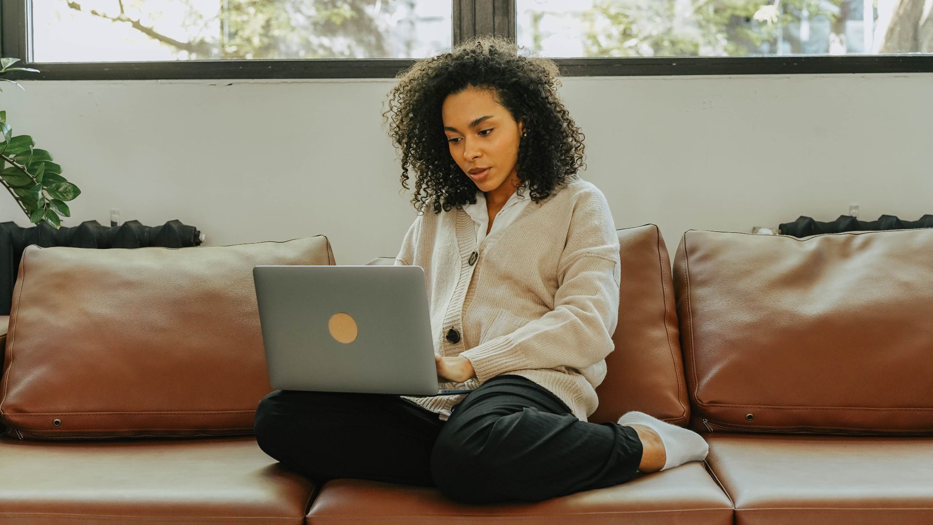 A woman with curly hair sits comfortably on a sofa, working on her laptop indoors.