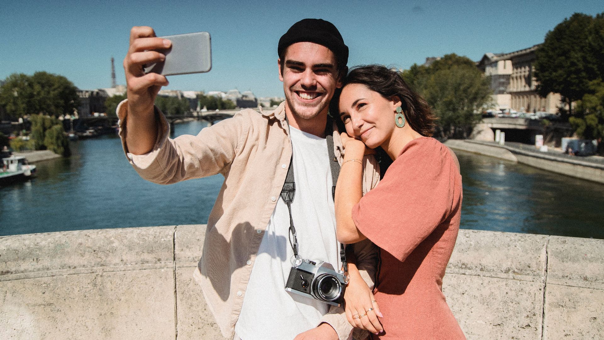 Young couple captures a romantic selfie on a bridge in Paris with scenic background.