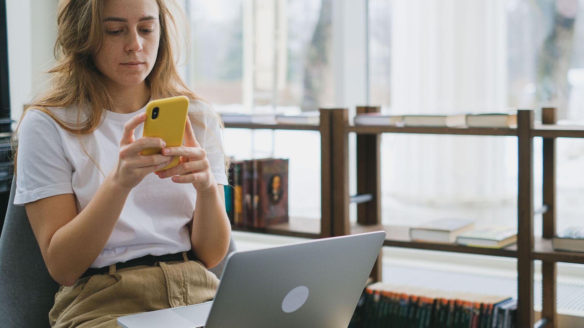 Young woman multitasking with smartphone and laptop in a cozy indoor setting.