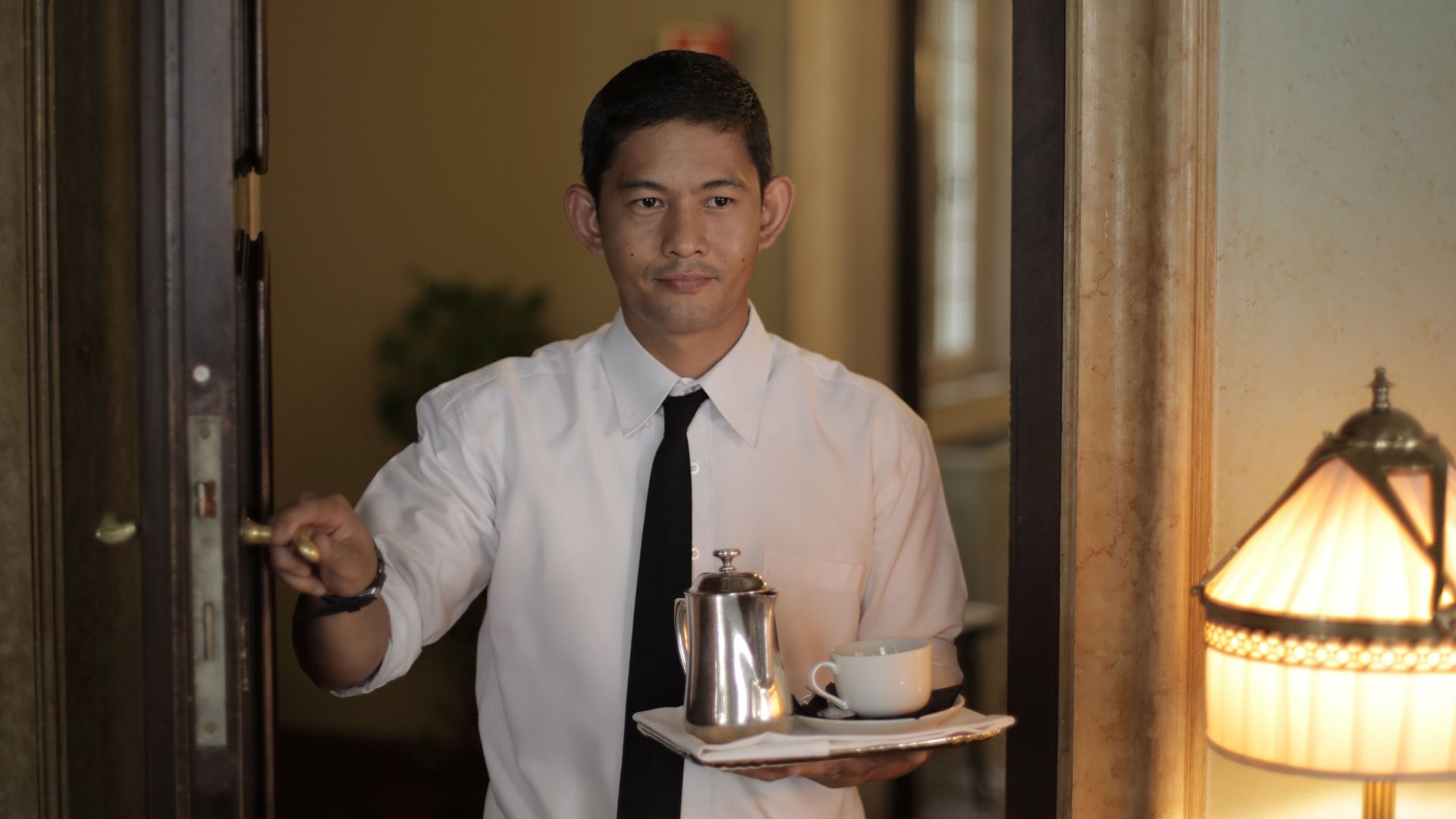 Positive ethnic male employee carrying tray with coffee while serving guest in cozy hotel room