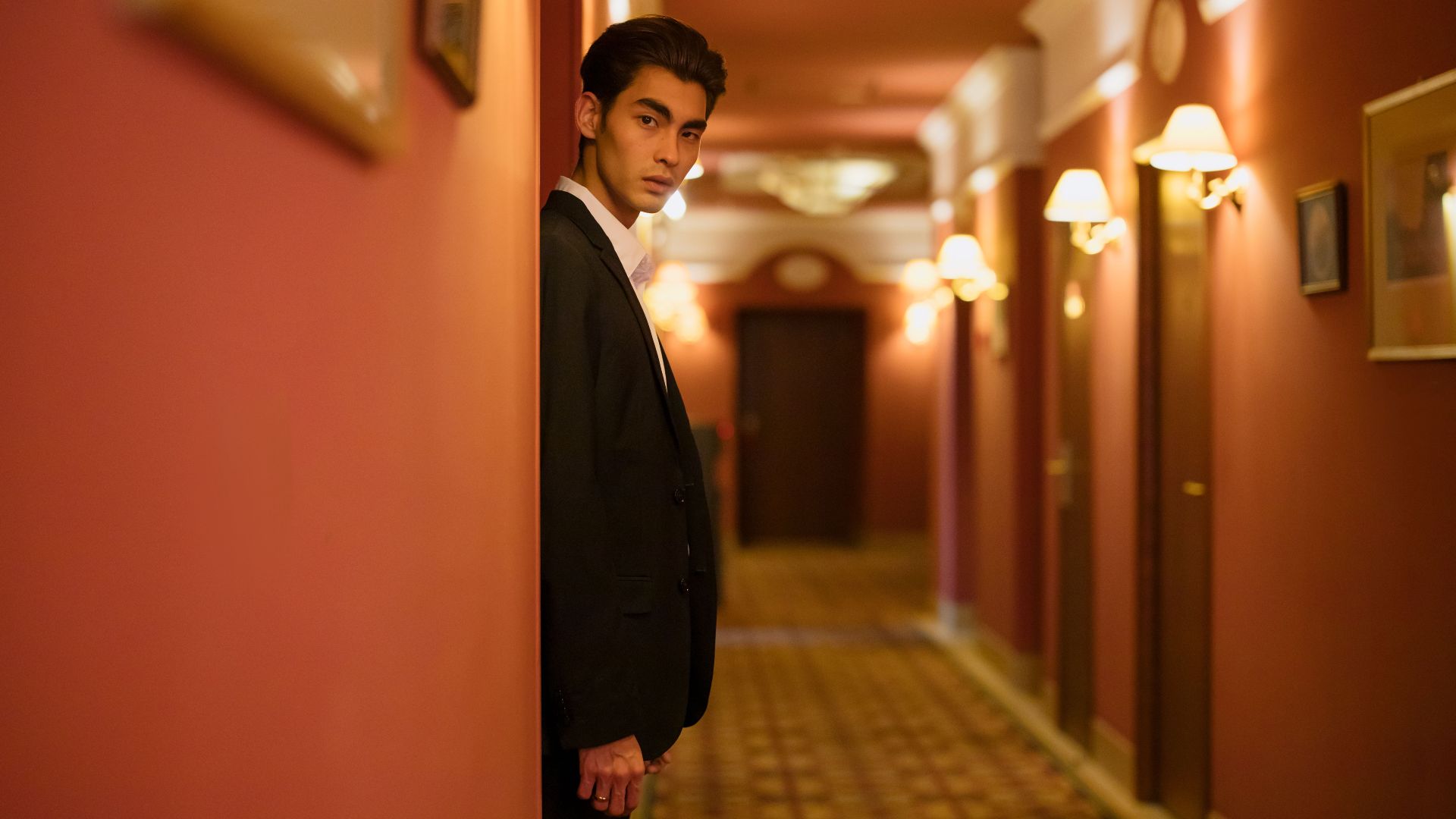 A fashionable man in a sleek black suit poses confidently in an elegant hotel hallway.