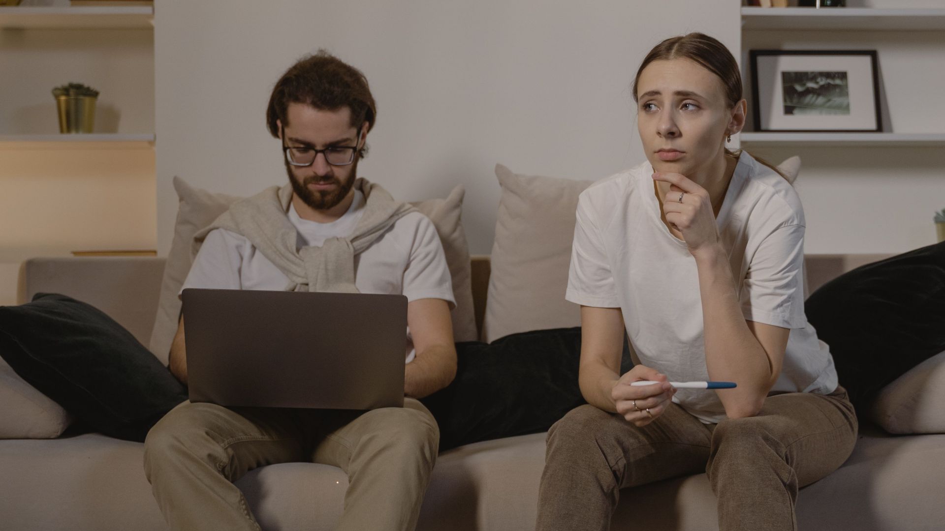 A couple sits on a sofa. The man is using a laptop, while the woman appears thoughtful.