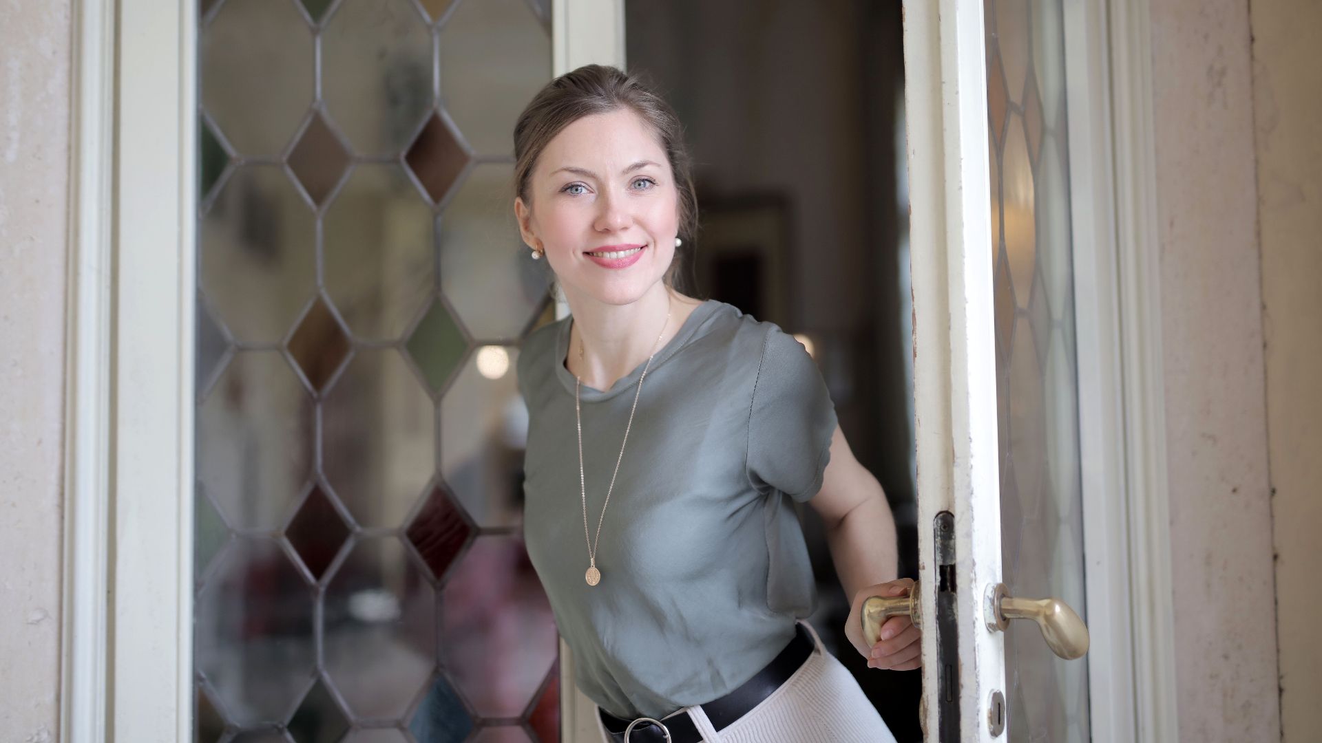 Young smiling female in casual outfit looking at camera and holding on doorknob while entering through stained glass vintage door in ancient building