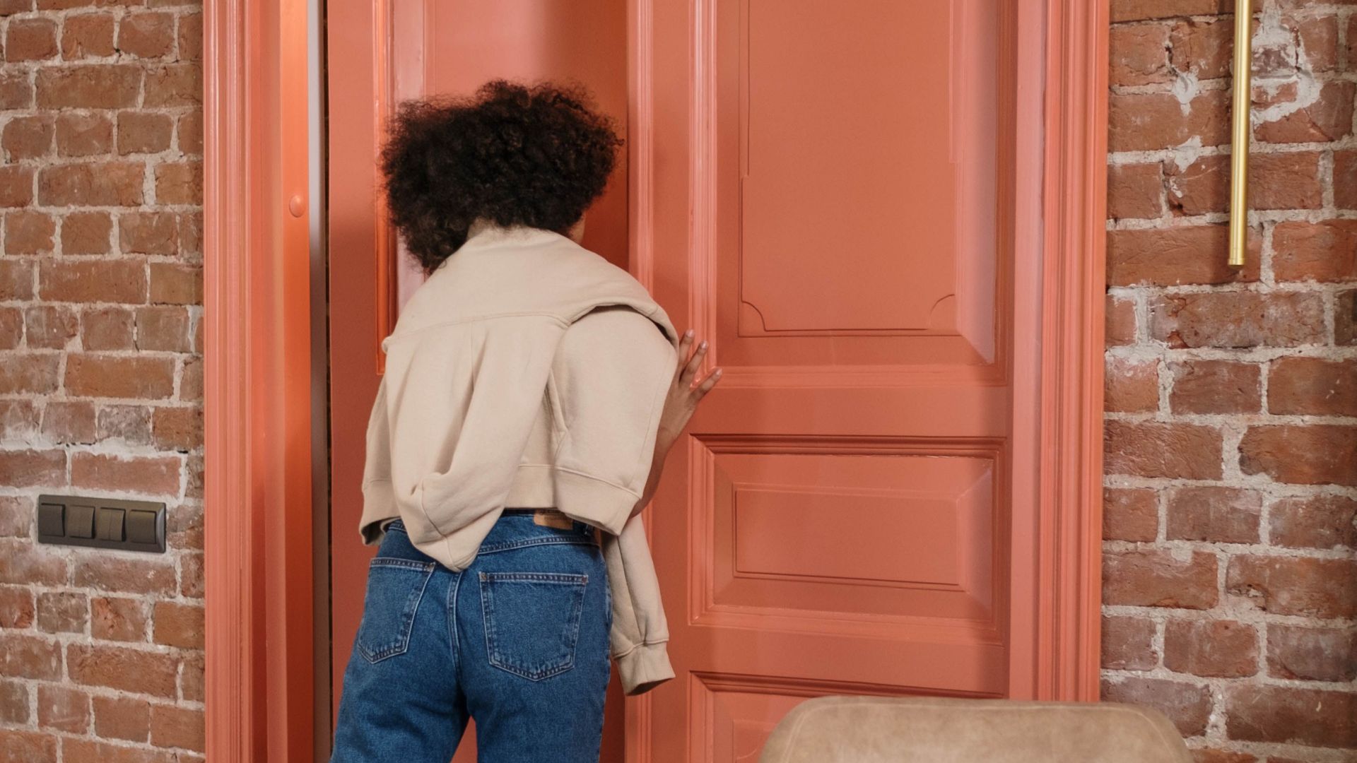 A woman with curly hair opens a large coral door in a modern loft room with a brick wall.