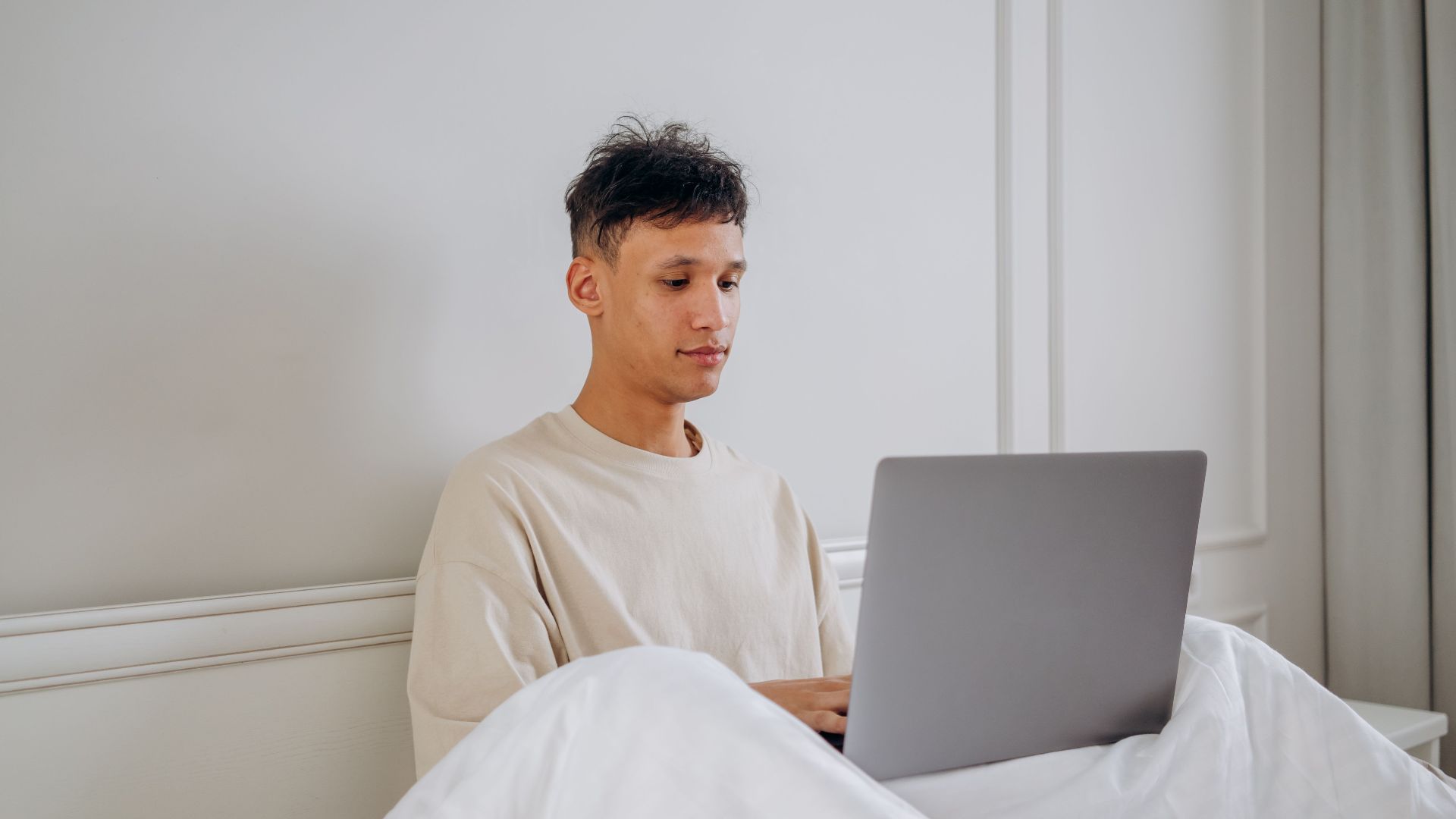 Young man sitting in bed using a laptop, embodying remote work lifestyle.