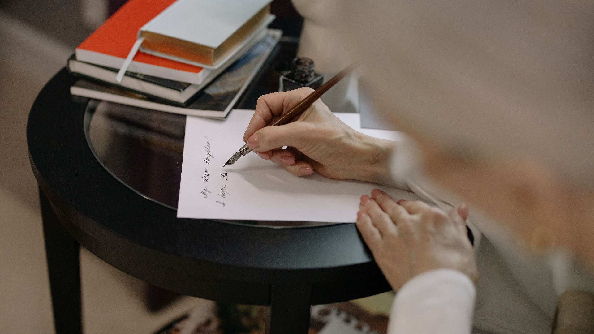 A person writes on white paper using a fountain pen, surrounded by books on a table.
