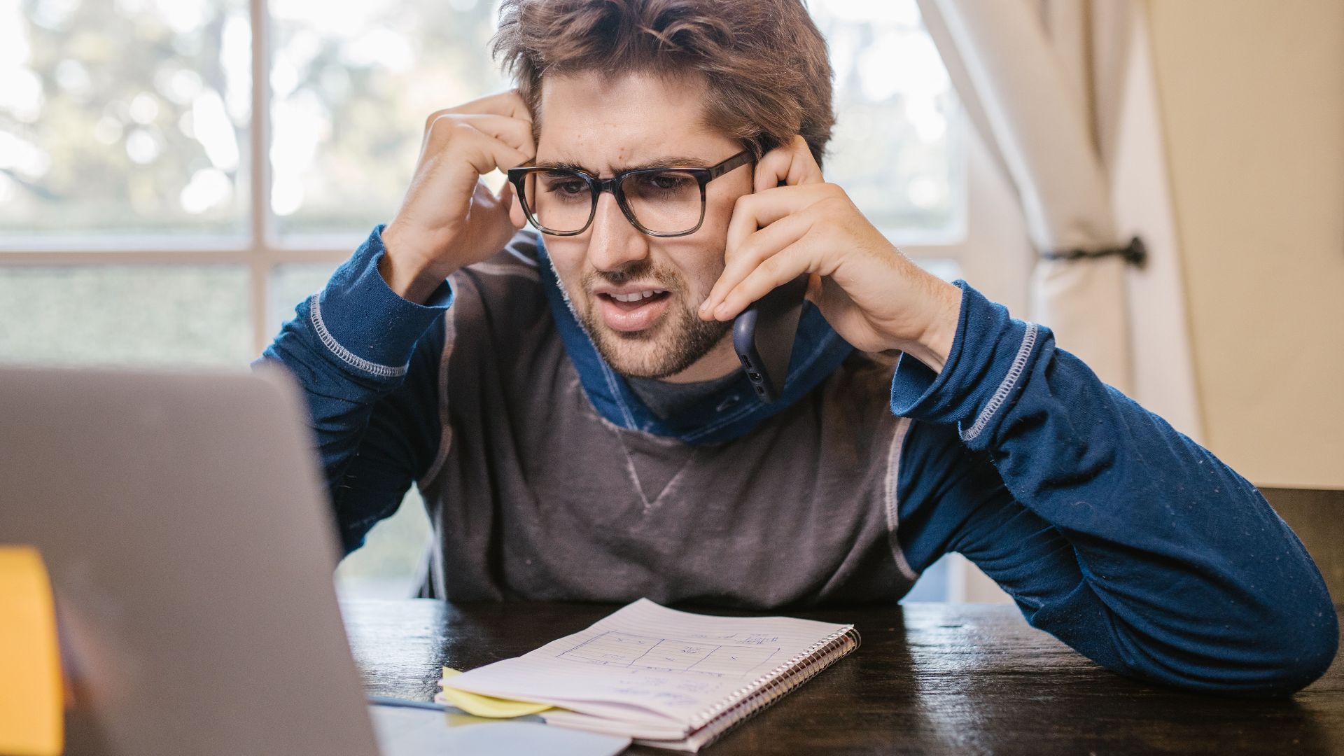 Man in glasses using laptop and smartphone at home, appearing confused.