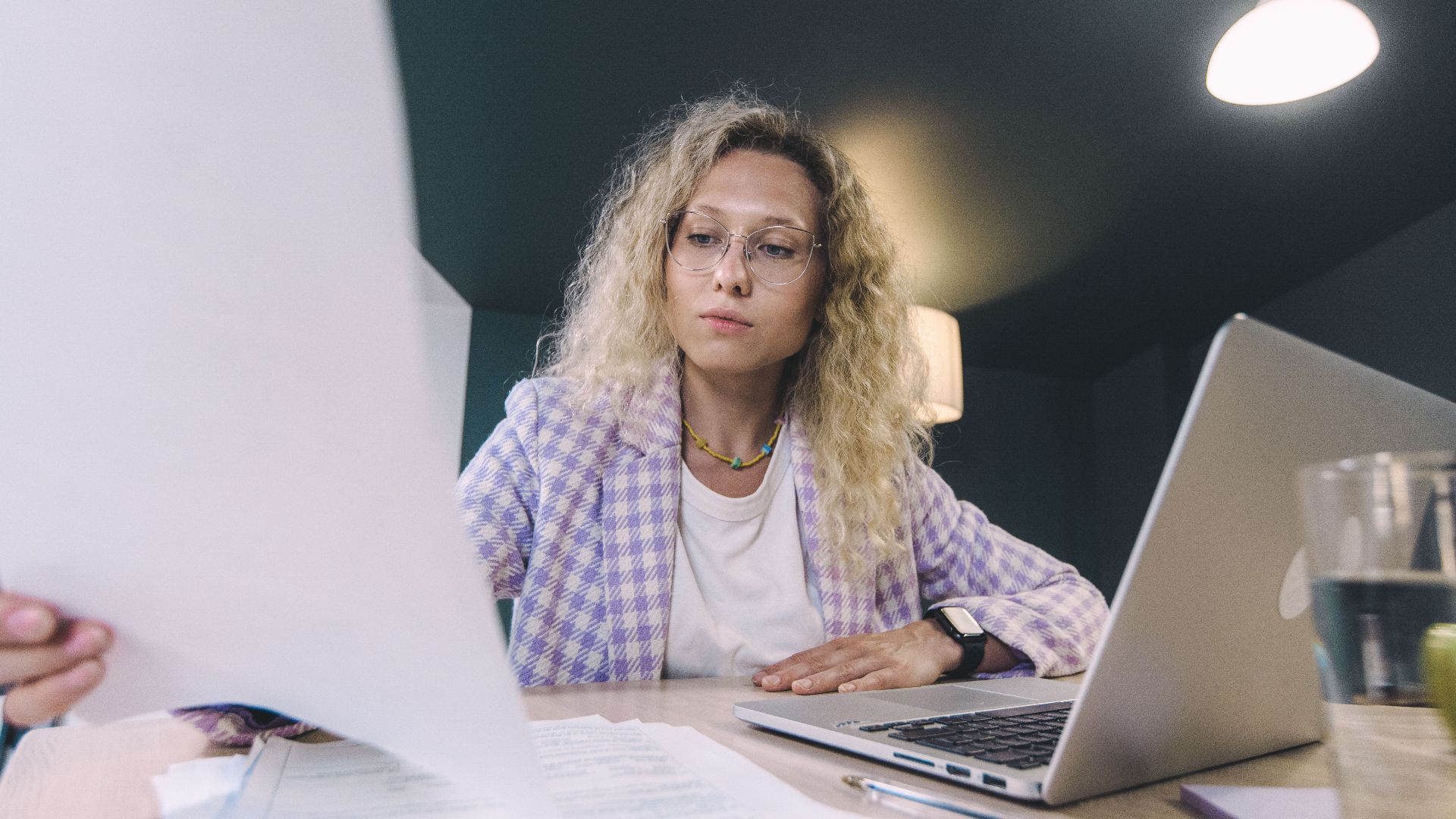 Woman in an office environment reviewing documents with focus, surrounded by technology.