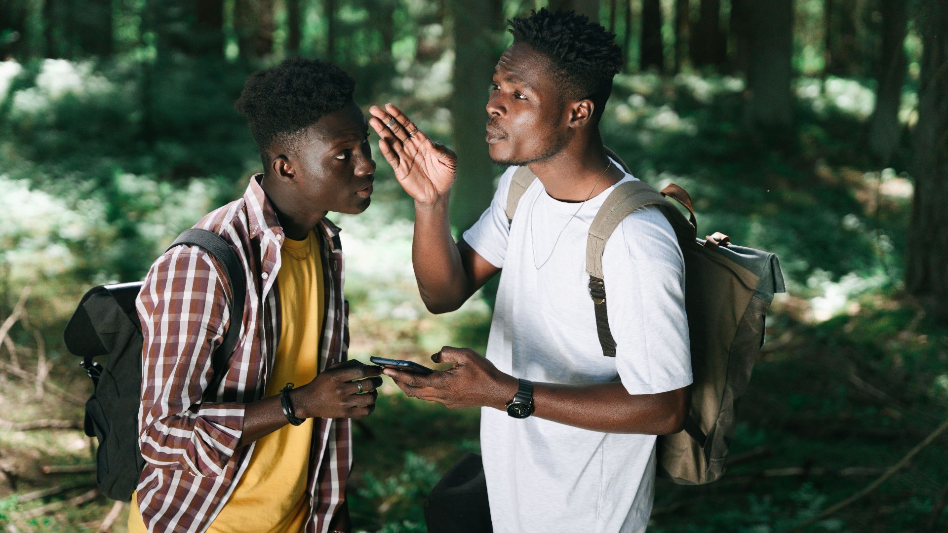 Two young men with backpacks discussing directions in a forest setting.