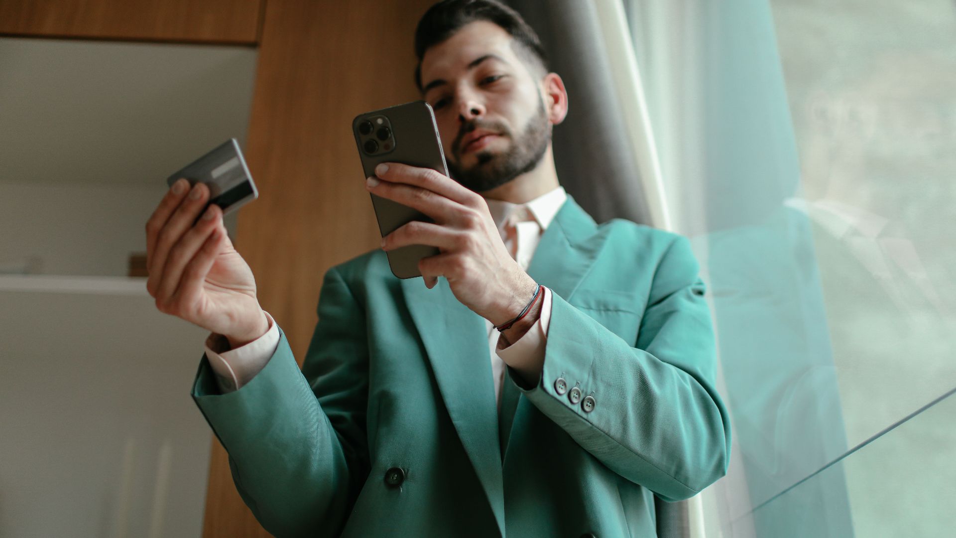 Man in teal blazer using smartphone and credit card for online shopping indoors.