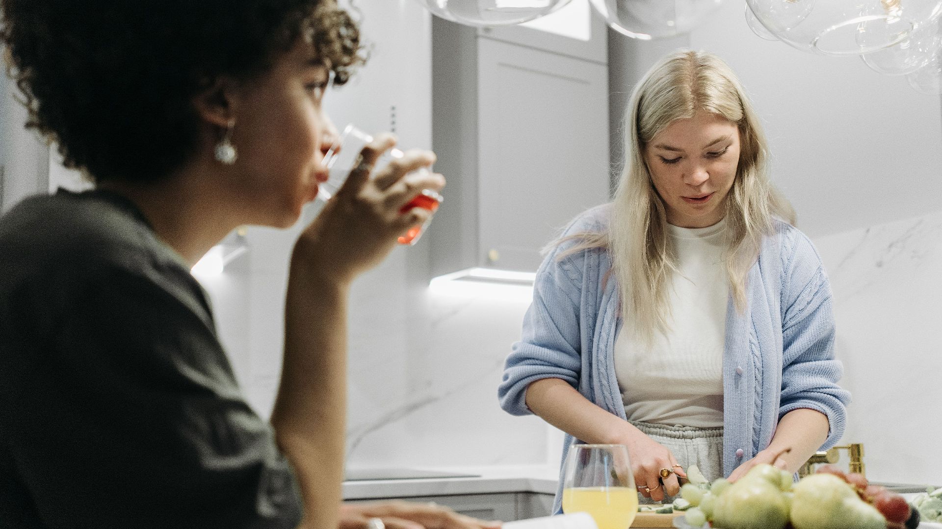 Two women in a modern kitchen; one slicing fruit, the other drinking. Bright and cozy setting.