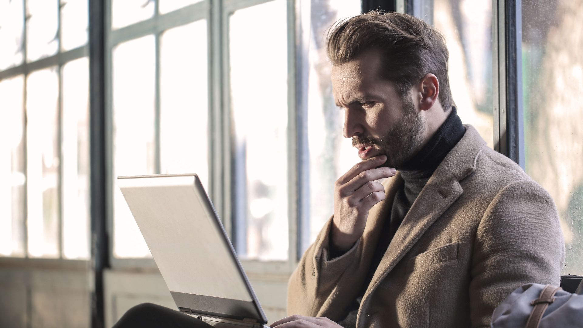 Man looking contemplative while working on a laptop in a well-lit indoor space.