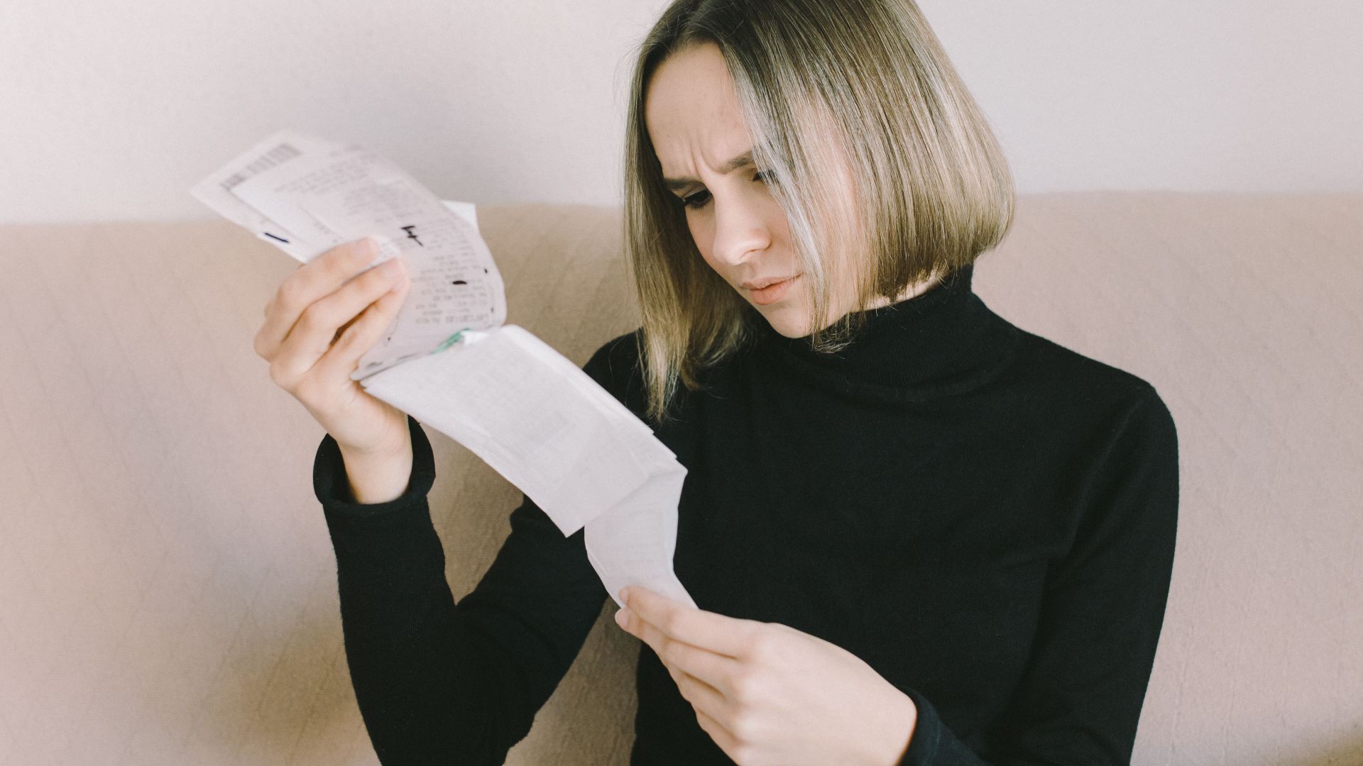 Concerned woman in black sweater examining bills on beige sofa indoors.