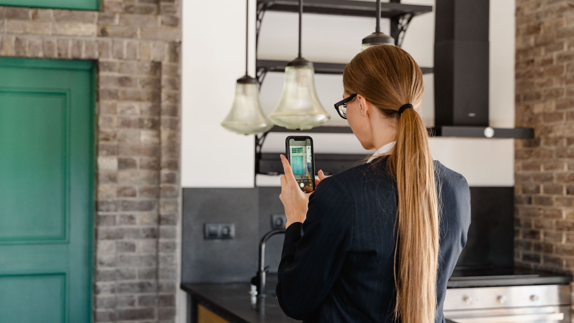 Rear view of a woman with long hair taking a photo with a smartphone in a stylish kitchen.