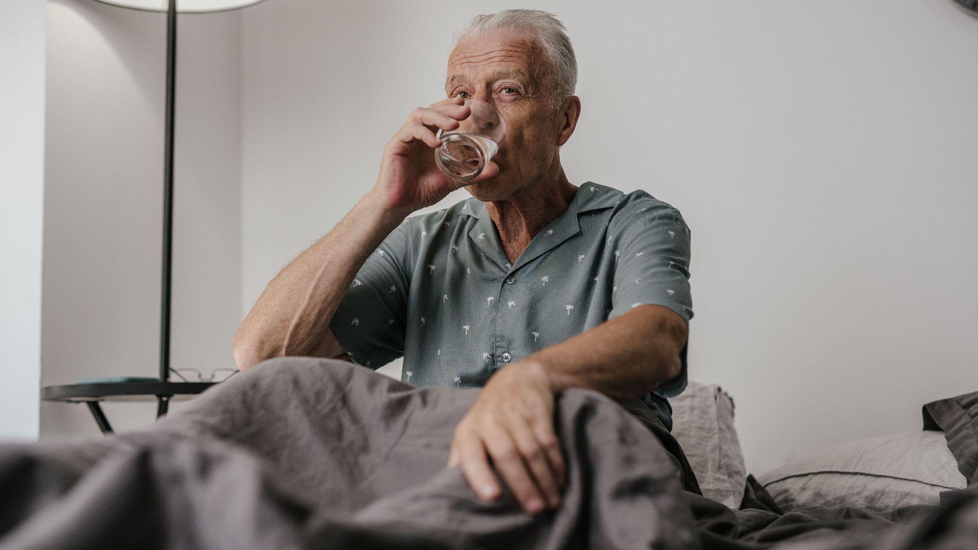 Senior man with gray hair drinks water in bedroom setting, conveying calm and routine.