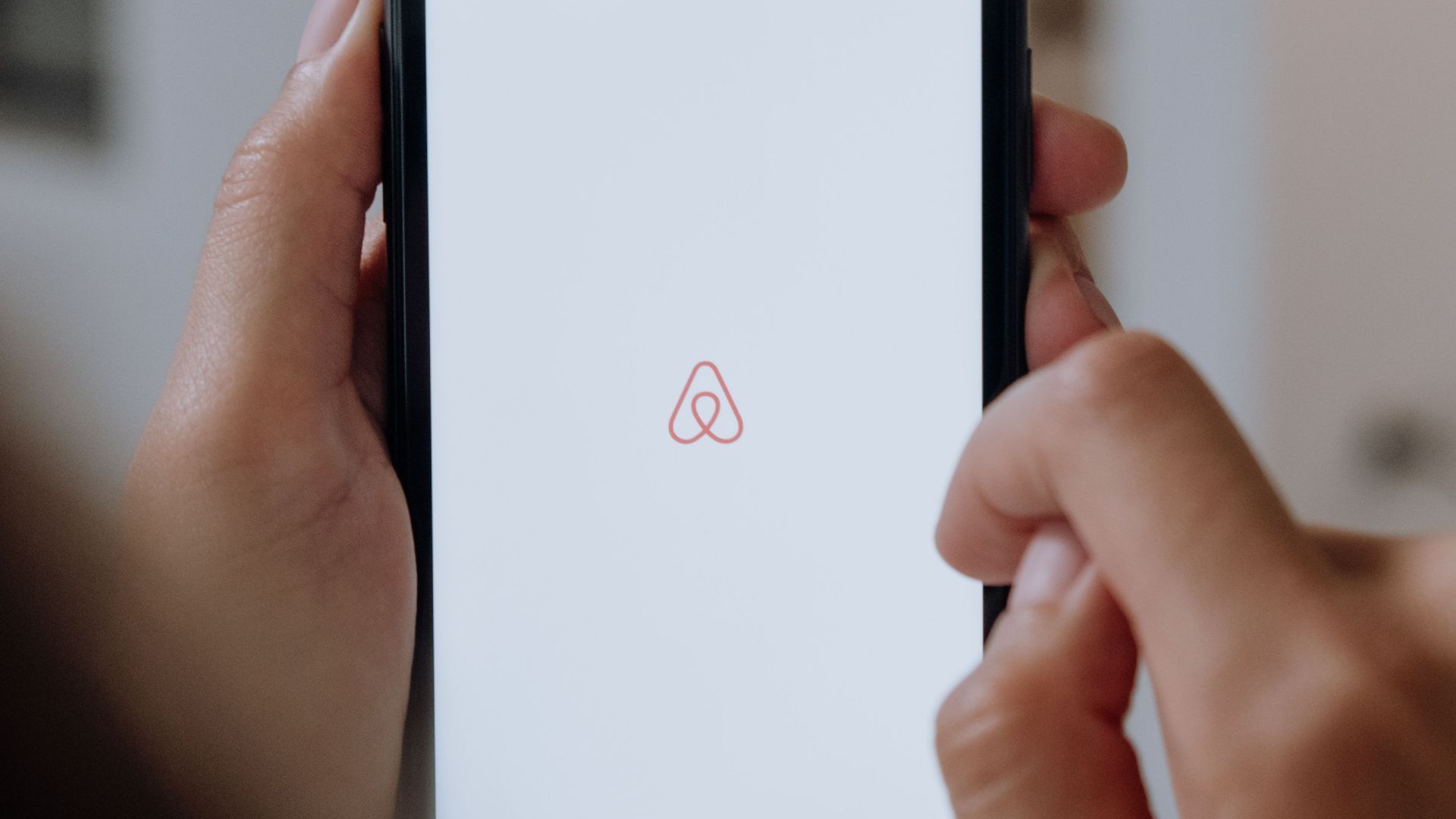 Close-up of hands holding a smartphone with Airbnb app logo on the screen indoors, highlighting modern technology.