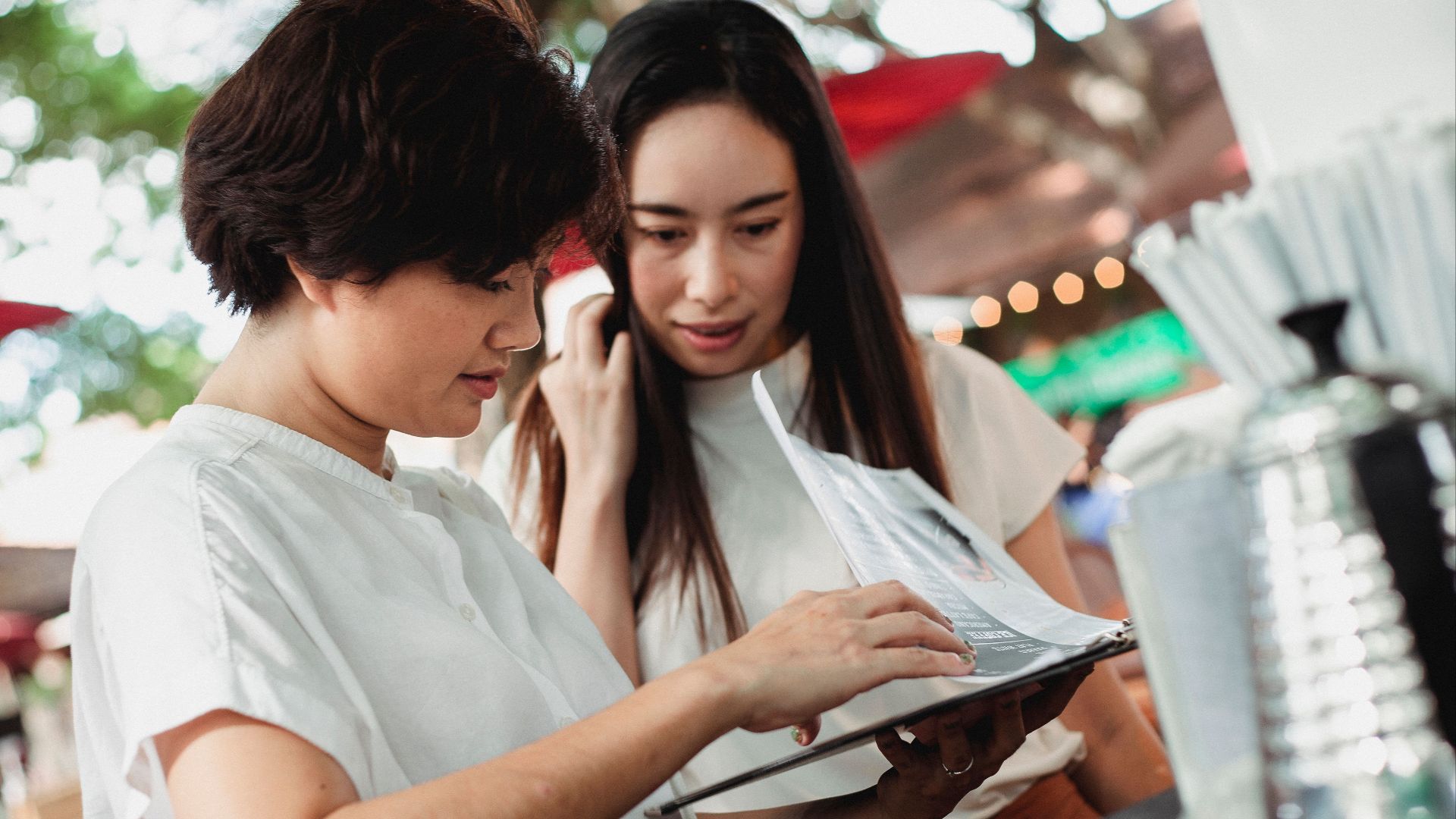 Glad young Asian females wearing white blouses reading menu while standing at street cafe counter on sunny weather