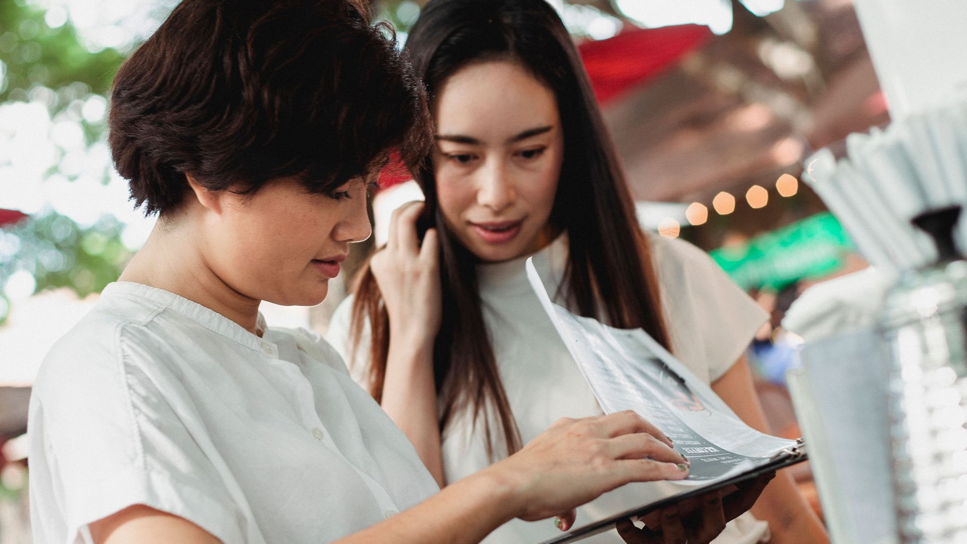 Glad young Asian females wearing white blouses reading menu while standing at street cafe counter on sunny weather