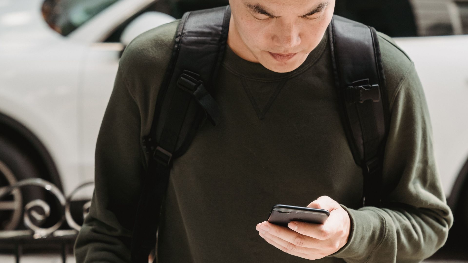 Asian deliveryman in beanie checks phone while carrying package outdoors.