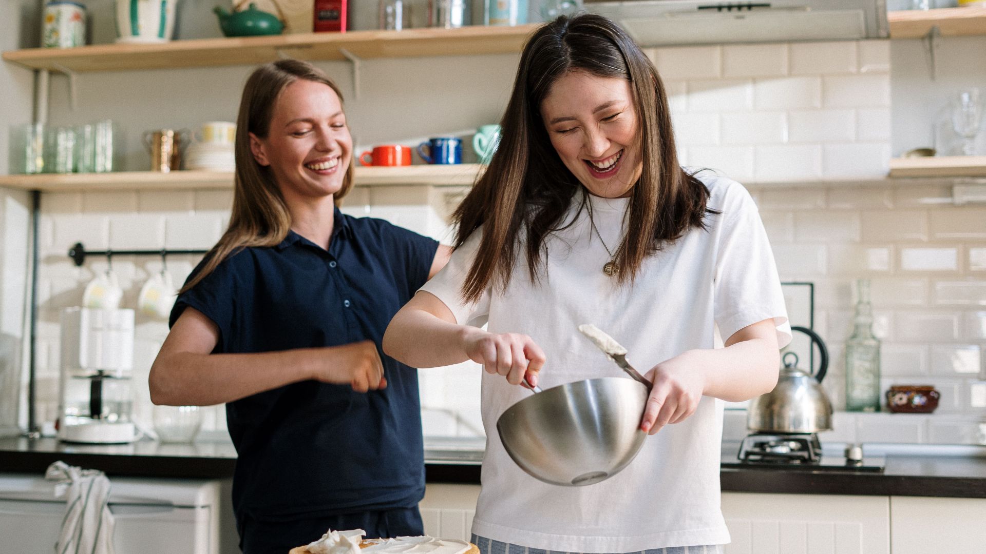 Two women cooking and laughing while preparing a cake in a cozy kitchen.