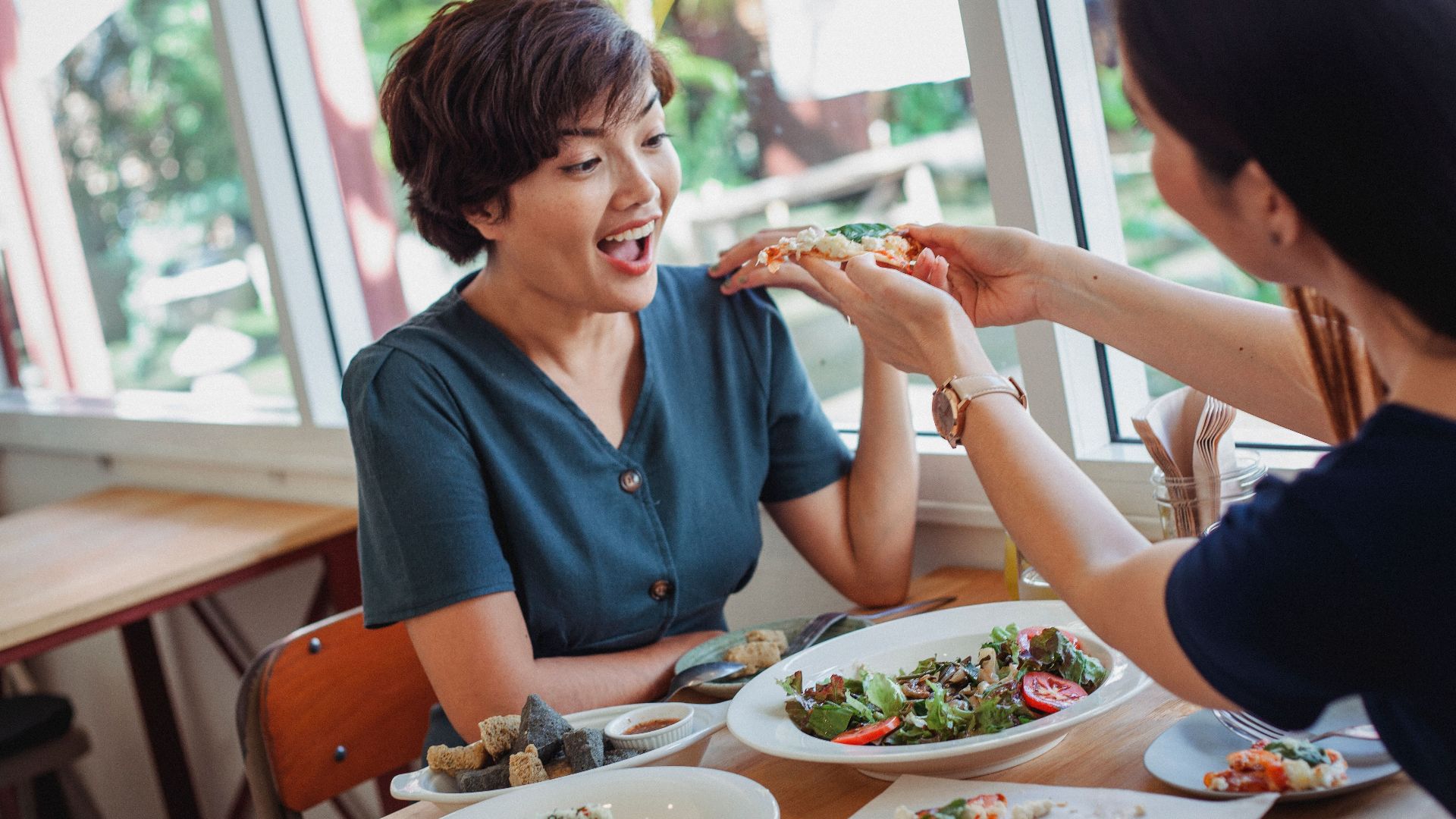 Unrecognizable woman giving delicious slice of pizza to exited Asian female while sitting in restaurant at table with tasty food