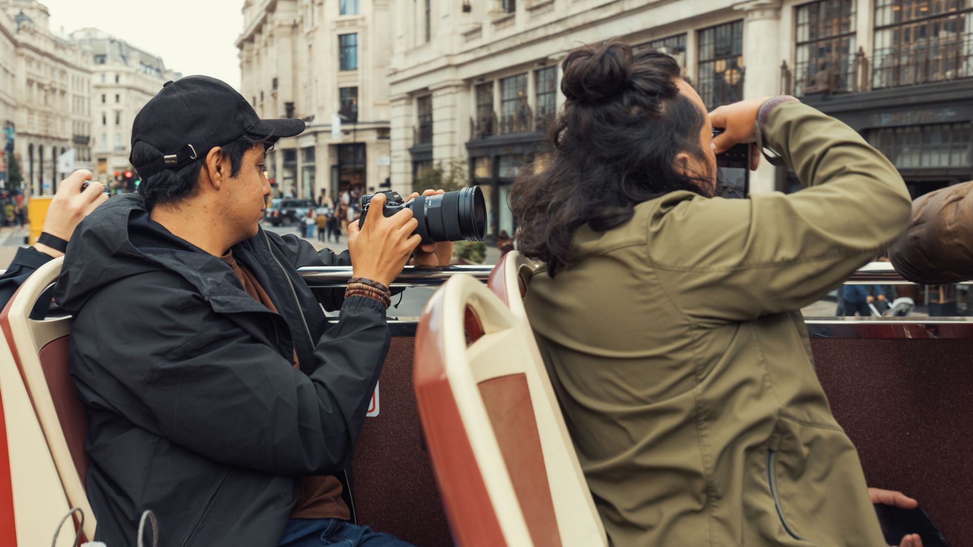 Travelers on an open top bus take photos of bustling city streets and architecture.
