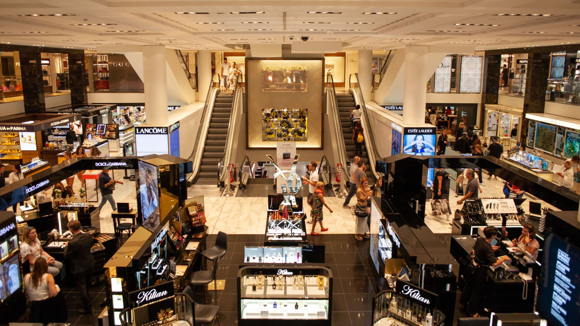 A bustling scene inside a modern shopping mall with escalators and various retail stalls.