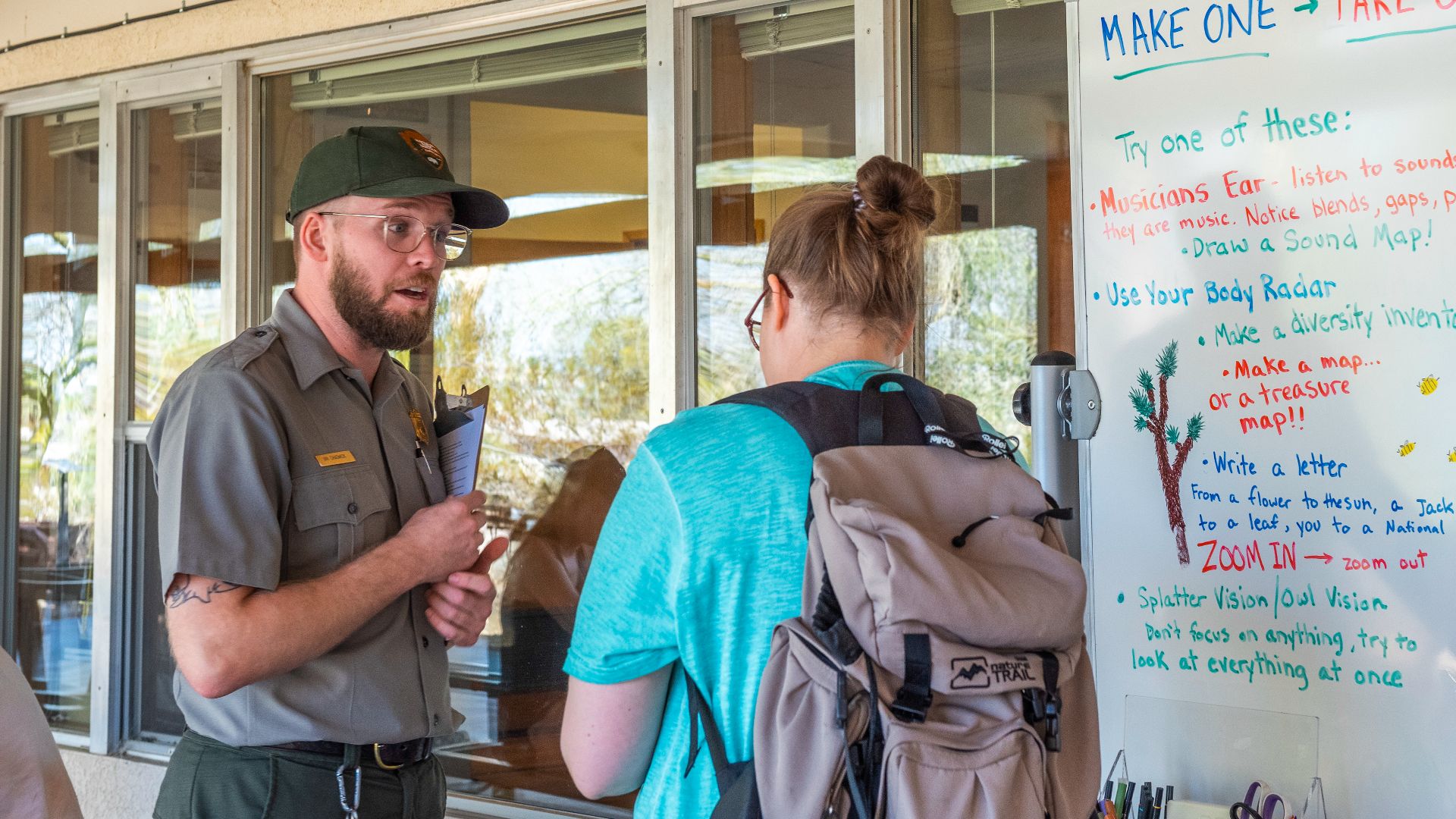 NPS / Emily Hassell

Alt text: A park ranger talks with a visitor outside of the Oasis of Mara Visitor Center in front of a table of drawing supplies.