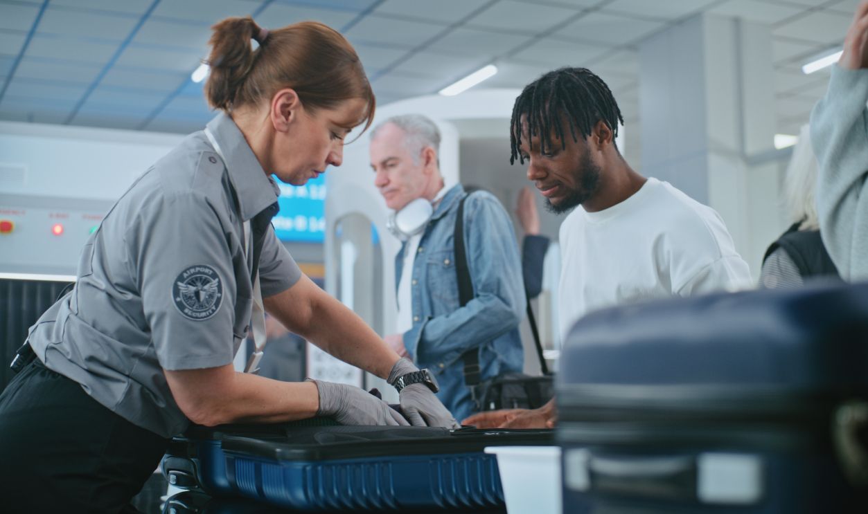 Security Checkpoint in Airport Terminal: Female TSA Worker Inspecting Baggage of Passenger before Boarding Flight, Finding and Confiscates Liquid. Queue of Diverse People During Screening Procedures.