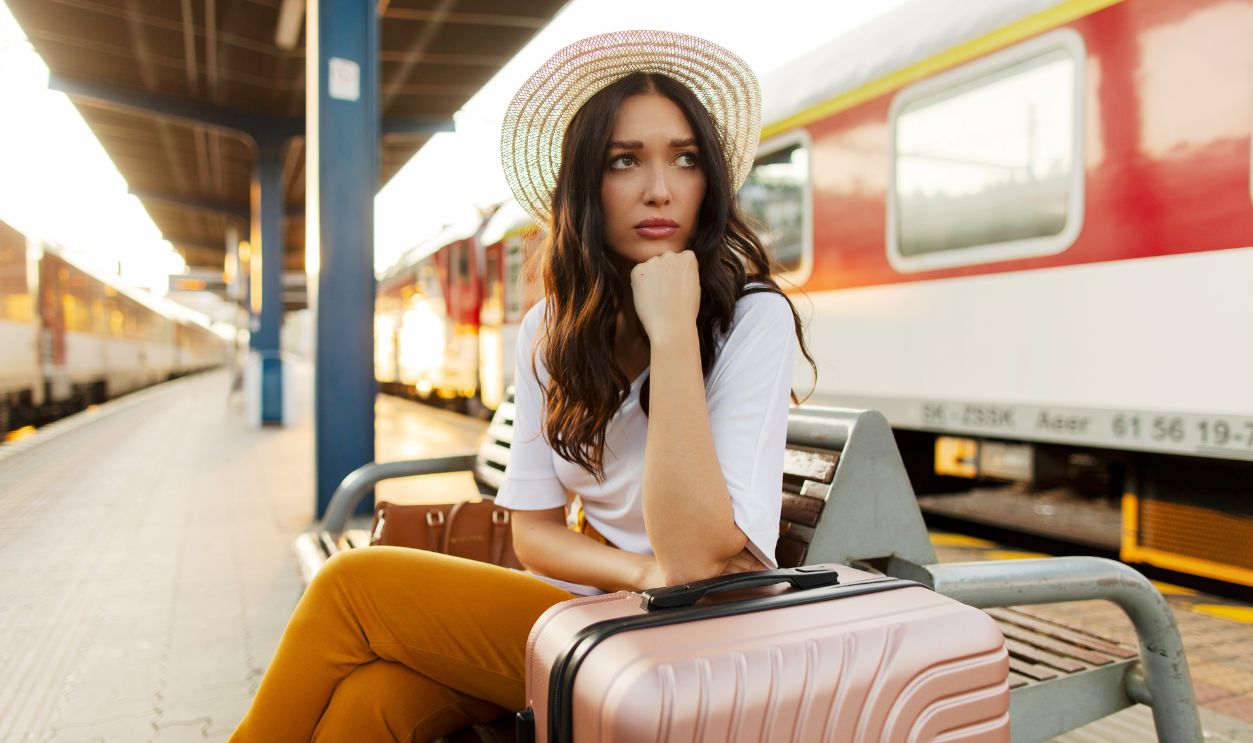 Despaired sad woman with suitcase being late for transport, sitting on bench at railway station platform