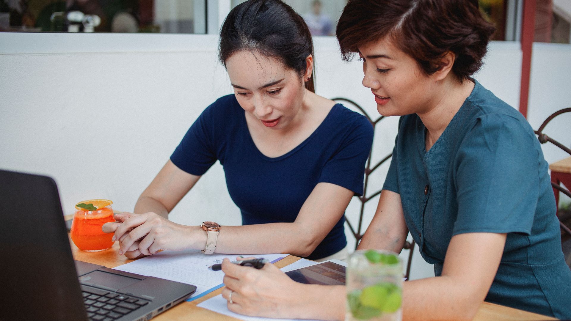 Concentrated female entrepreneurs in casual clothes sitting at table with gadgets and cocktails while signing documents