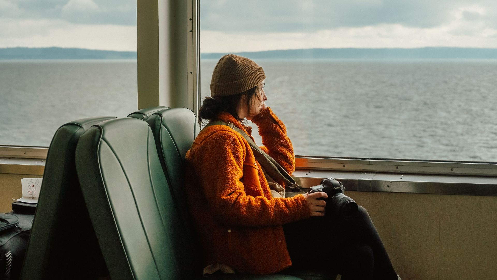 Woman wearing beanie gazes out ferry window with a camera in hand, capturing serene moments.