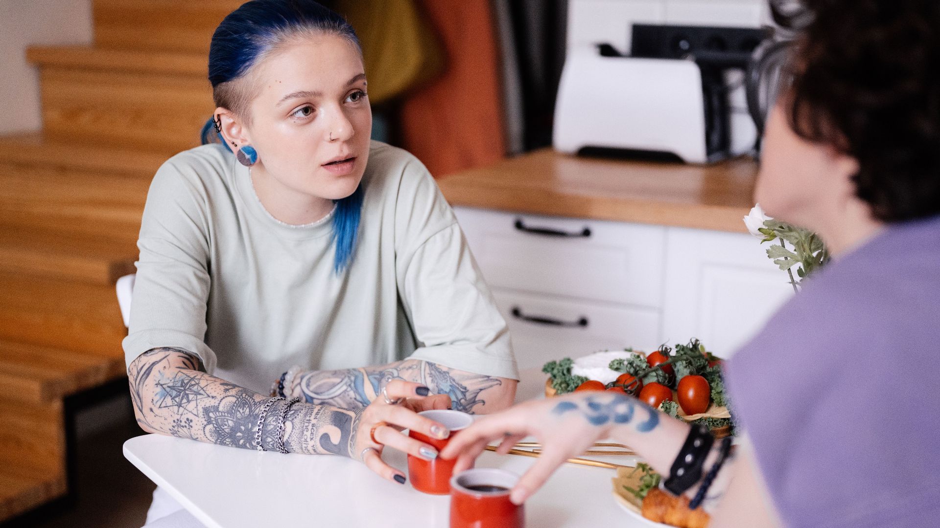 Two tattooed women with dyed hair enjoy coffee at a table indoors, chatting intimately.