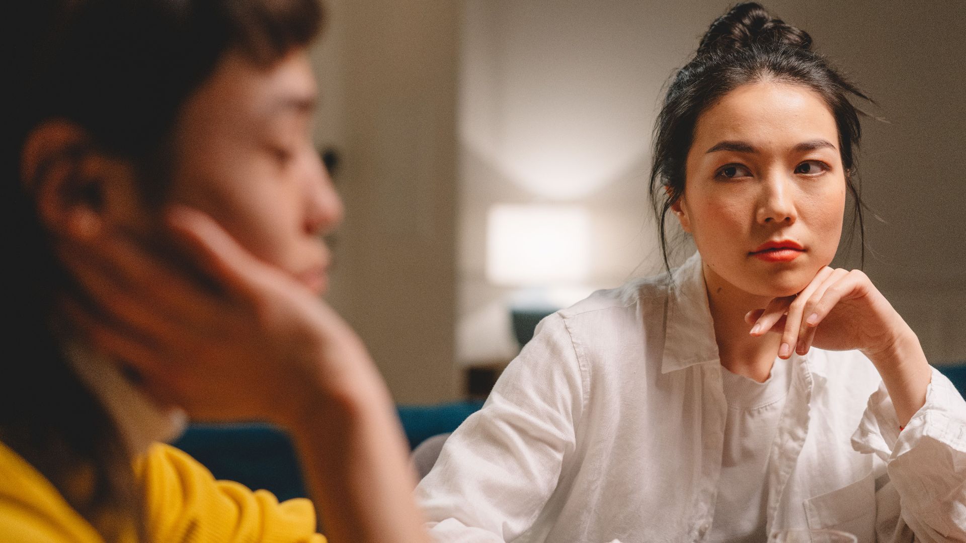 Serious concentrated young Asian female friends with dark hair in casual clothes sitting at table with hands at chins and discussing problems at home