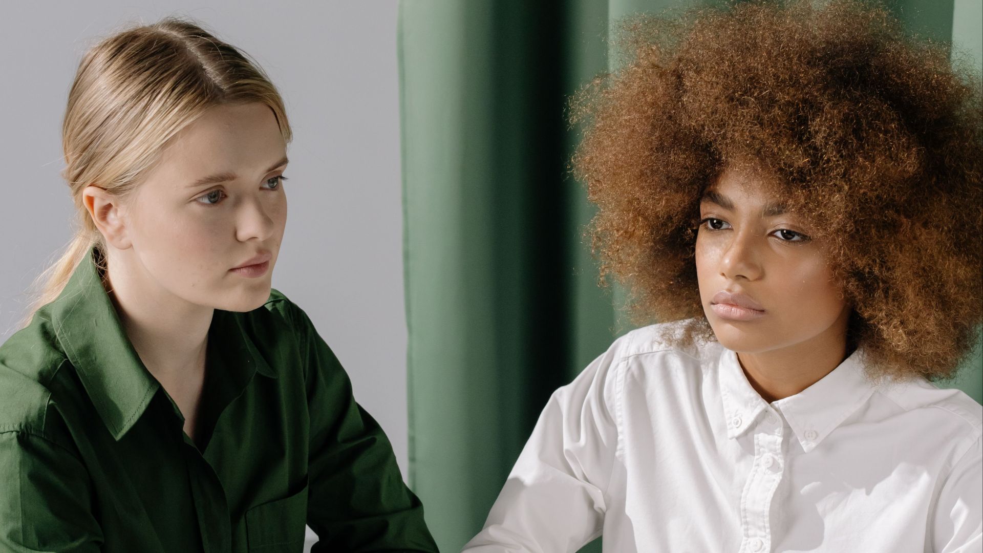 Two women, one Black and one Caucasian, sitting together with a serious expression in a minimalist indoor setting.