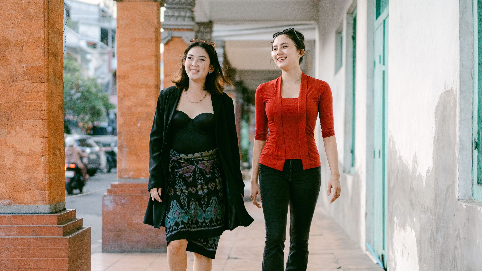 Two smiling women walk down an urban sidewalk, enjoying a sunny day in the city.