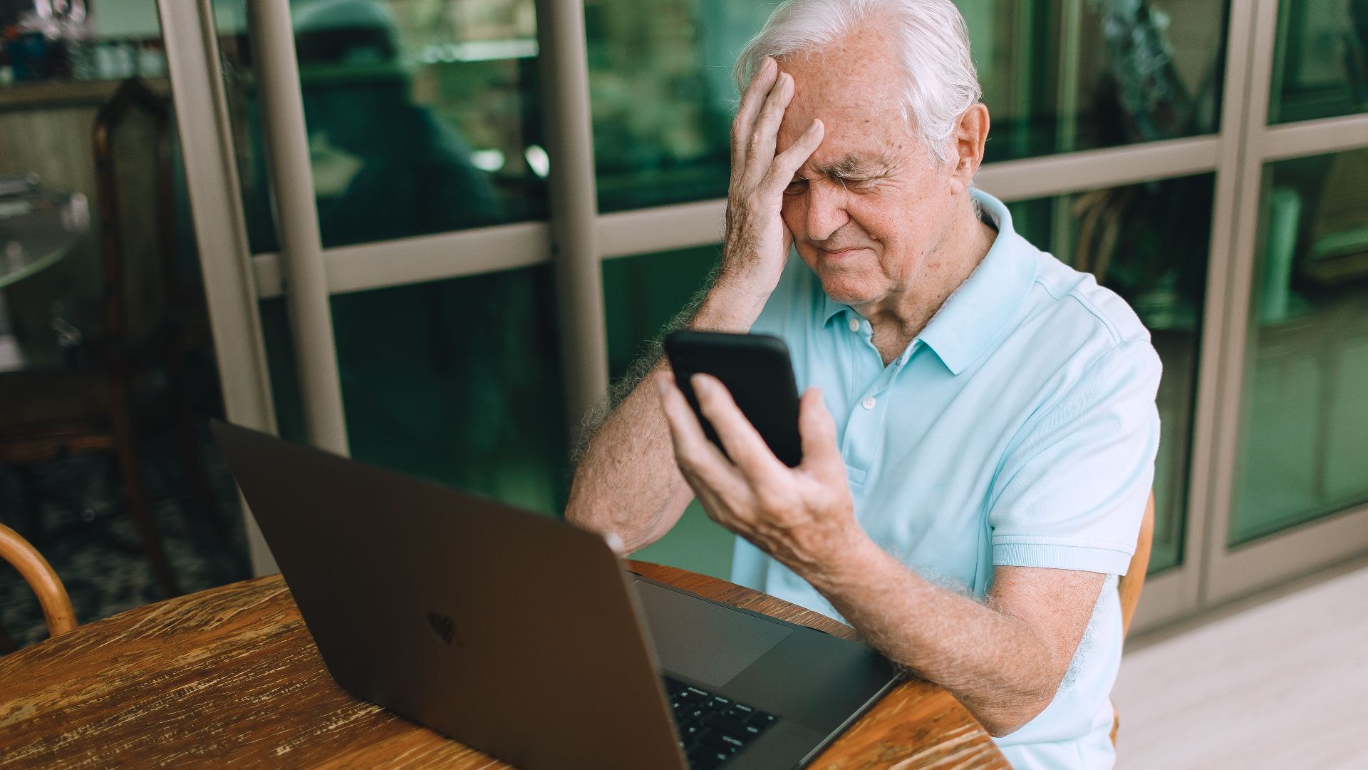 Elderly man frustrated by smartphone and laptop technology indoors at home.