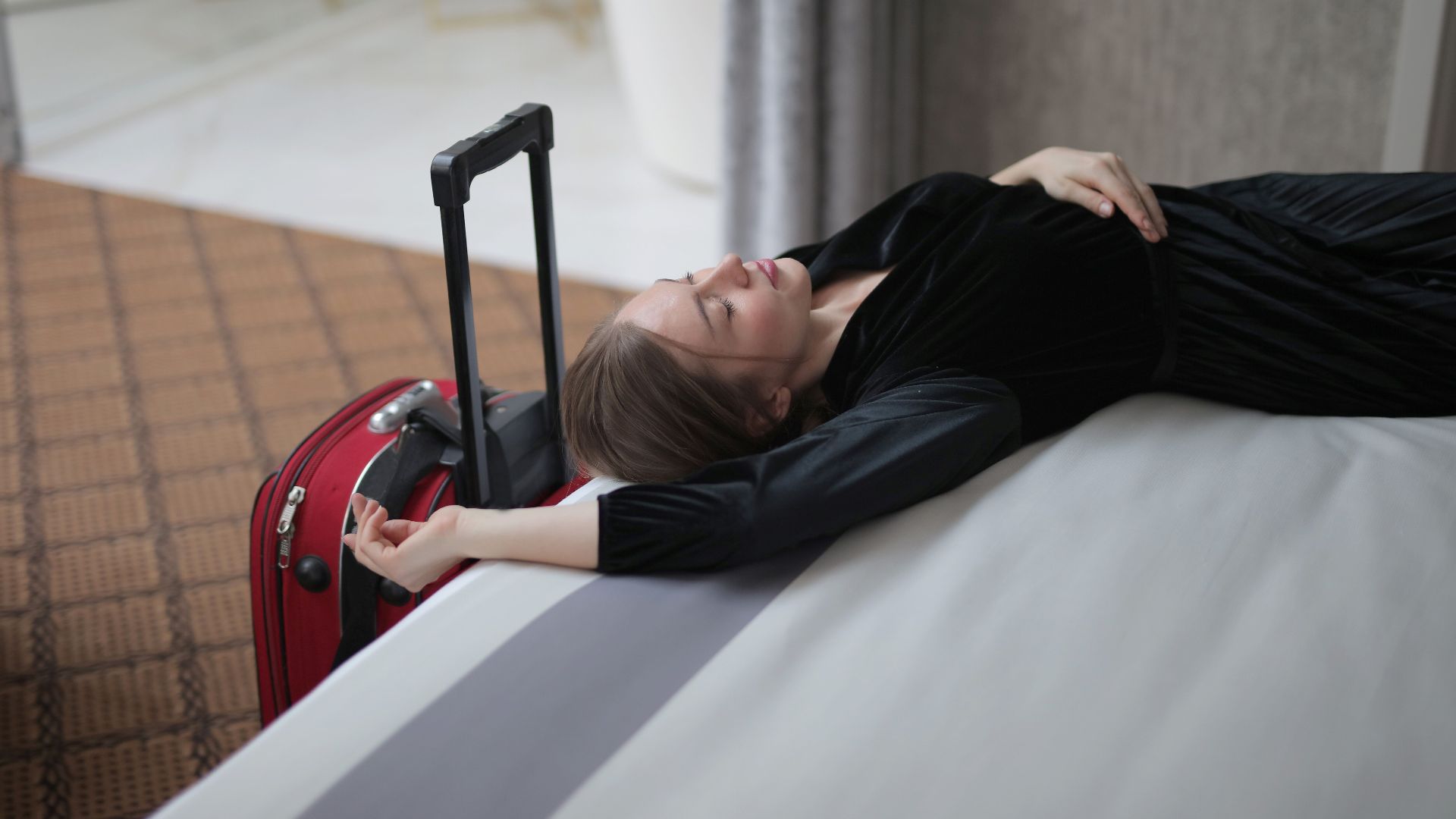 A woman resting on a hotel bed beside her suitcase, capturing travel fatigue.