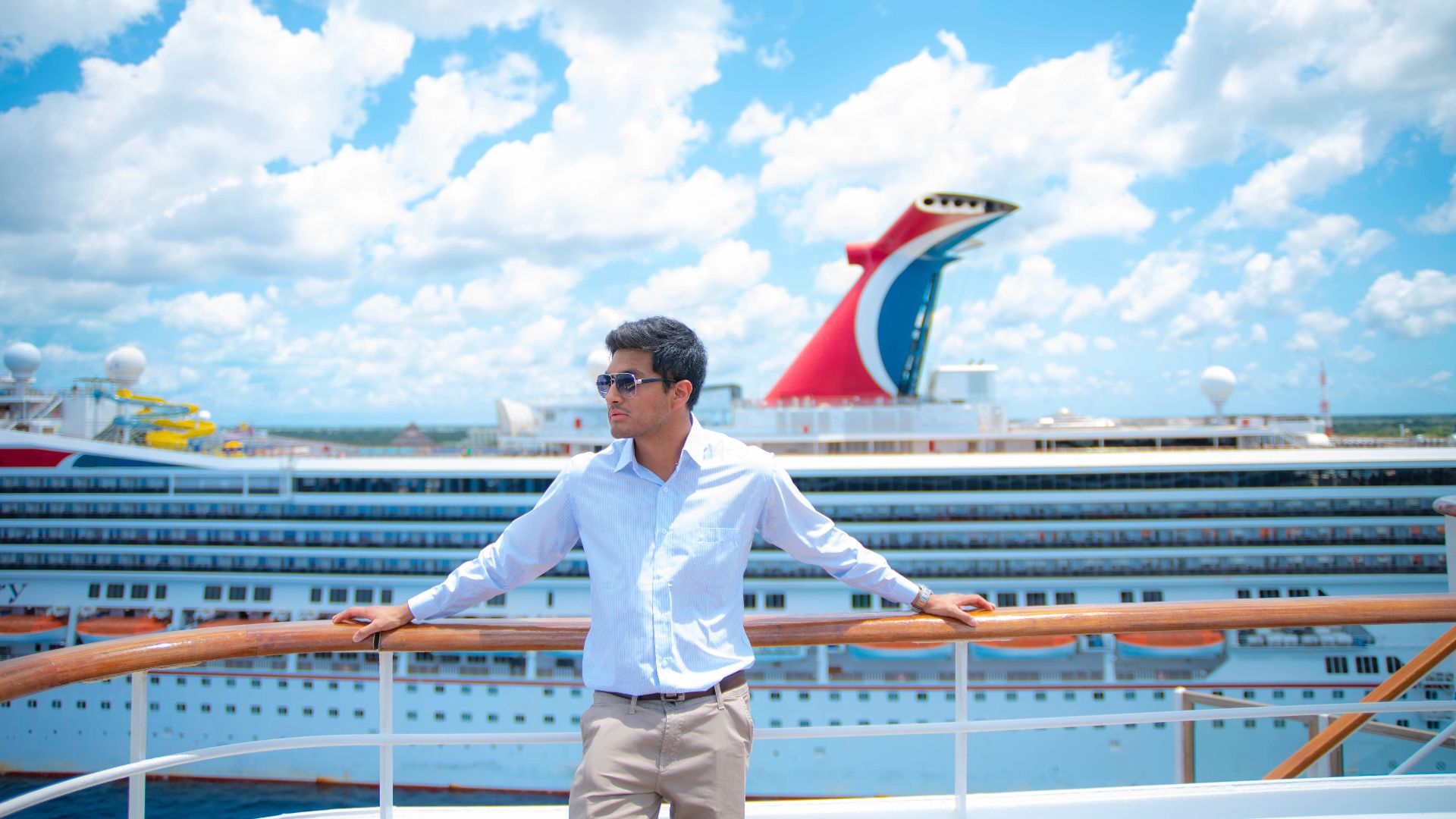 A man in sunglasses and white shirt enjoys a sunny day on a cruise ship deck with puffy clouds overhead.