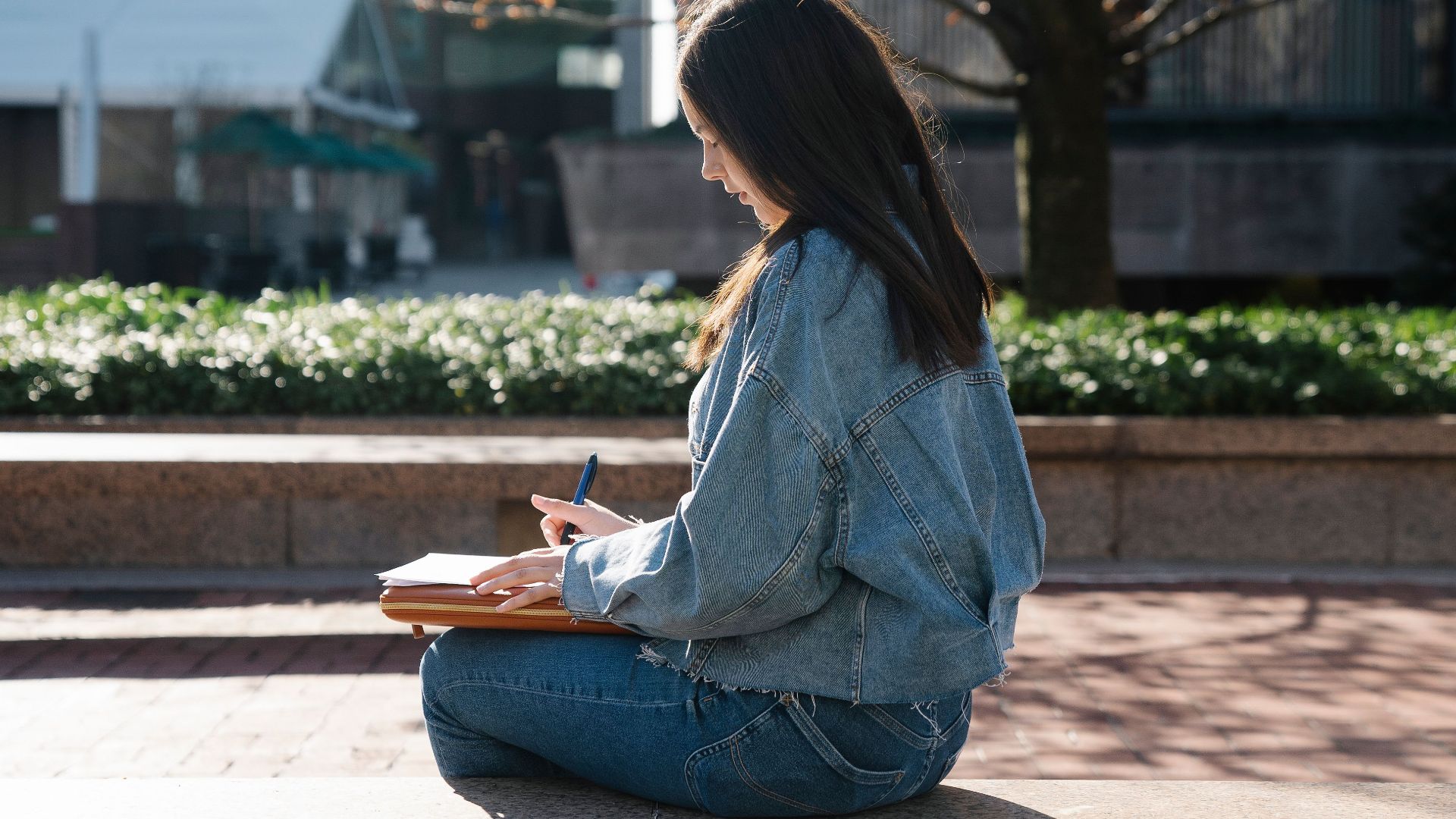 A young woman in casual attire writes in a notebook while sitting on a ledge in a park during a sunny day.