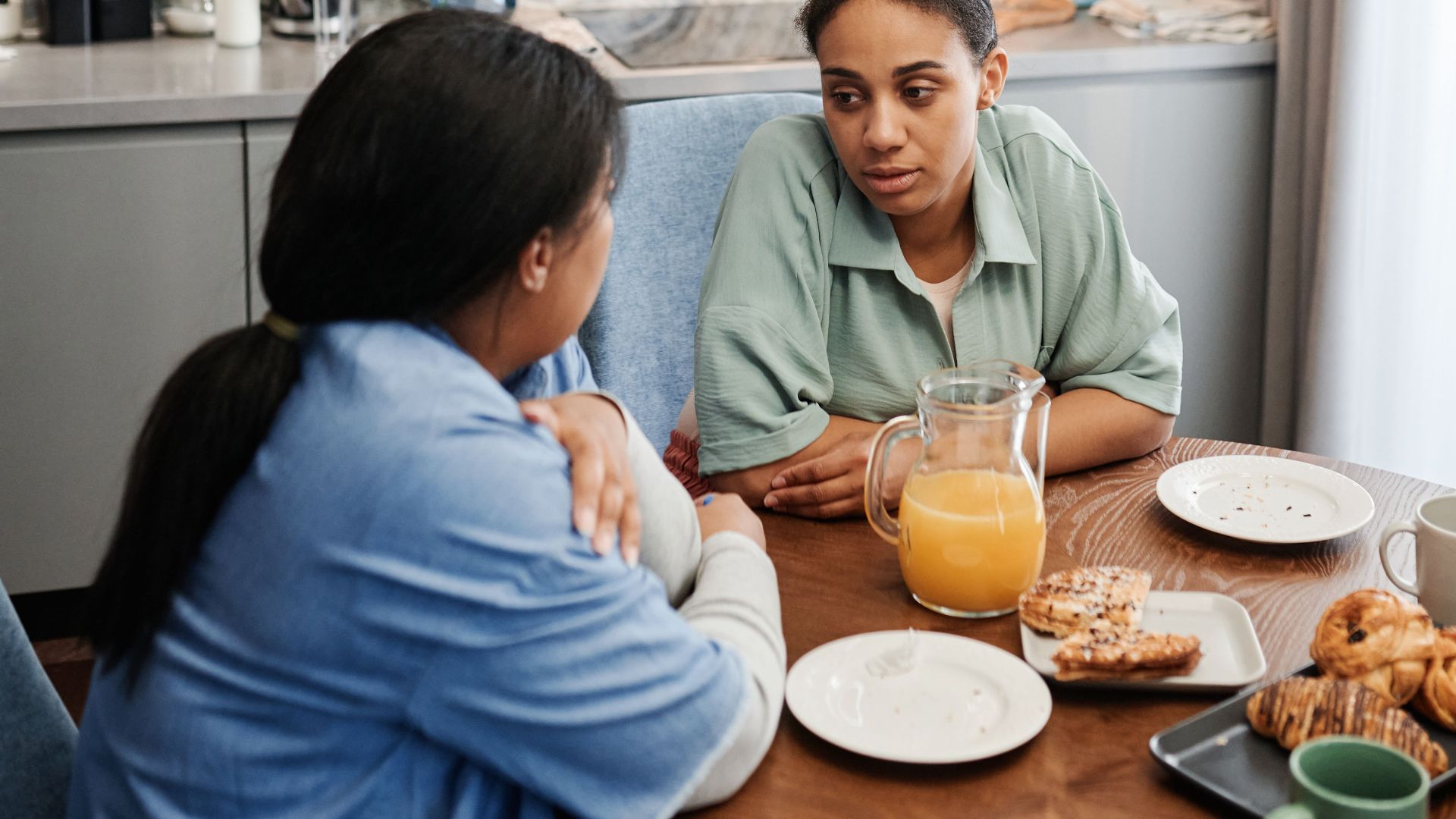 Two women engaged in a conversation at a kitchen table with breakfast. Modern interior setting.