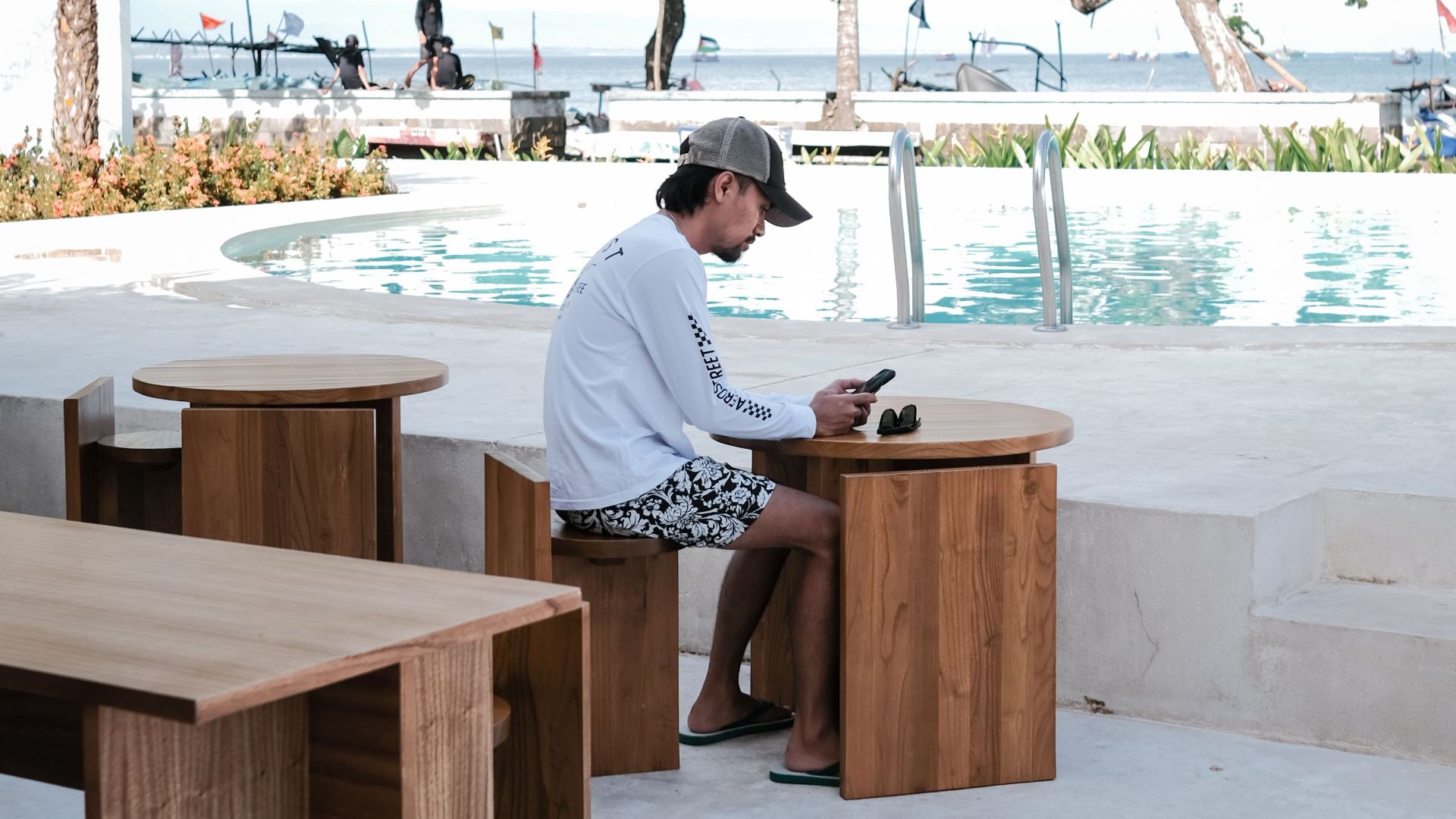 Man enjoying leisure time by the poolside in a tropical setting with wooden seating.