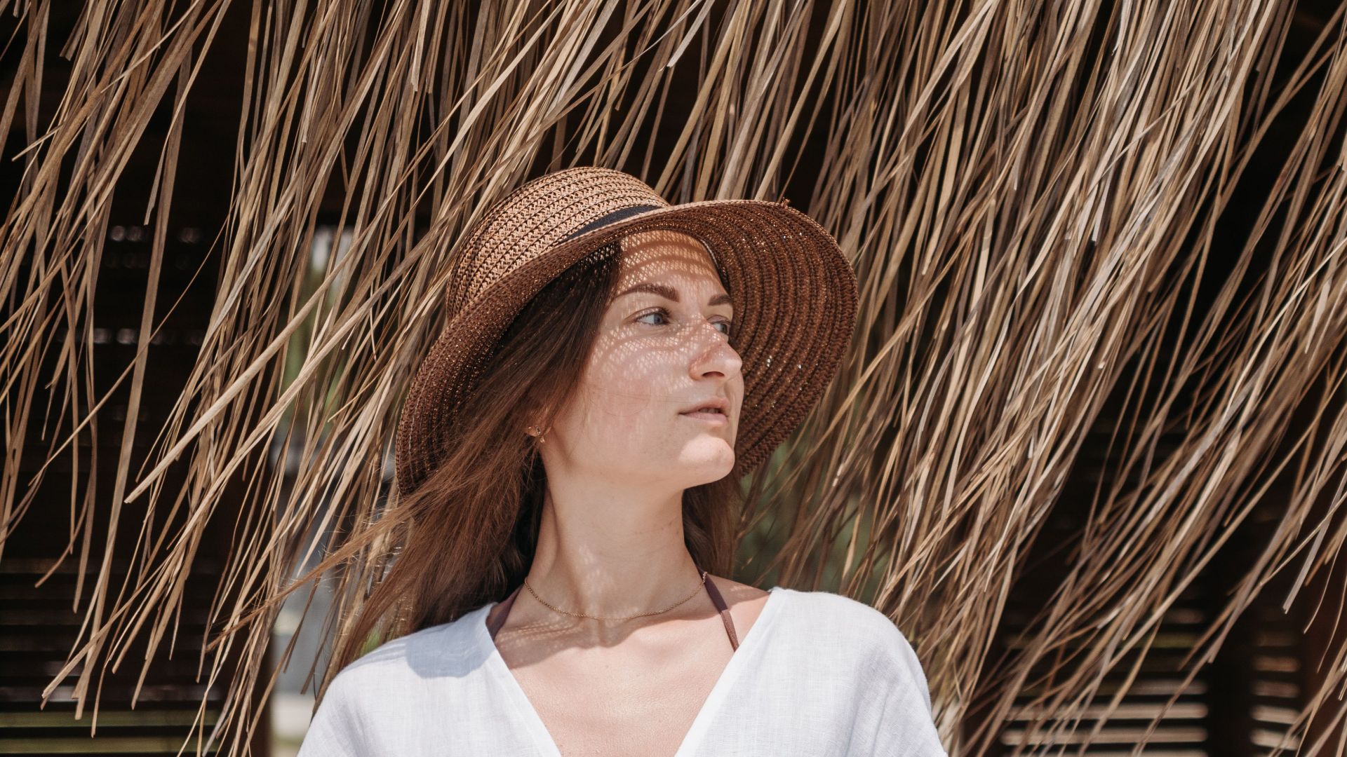 Woman in a white dress and straw hat standing under a thatched umbrella.