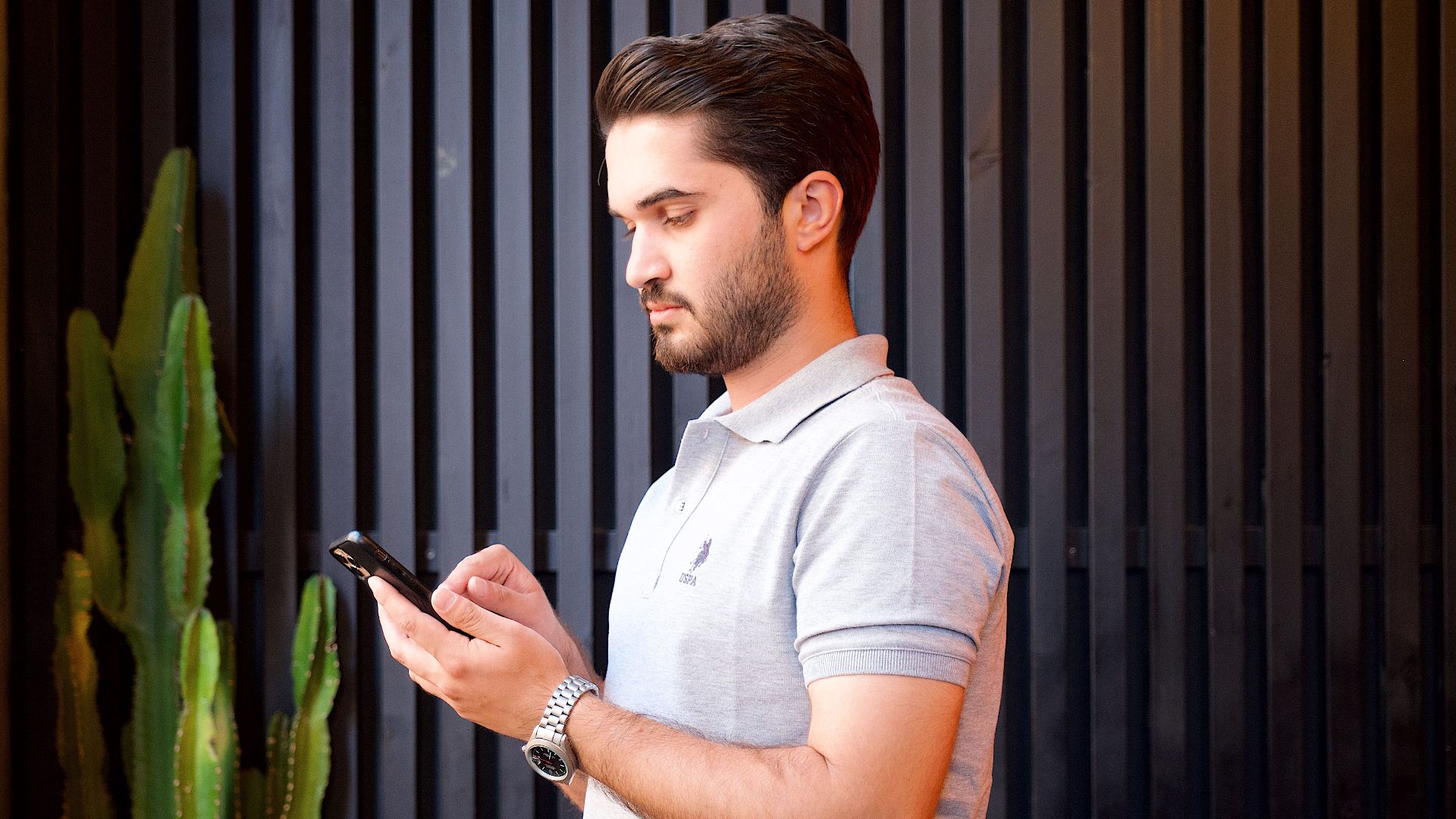 A young man in a casual polo engrossed in his mobile phone, standing by a cactus.