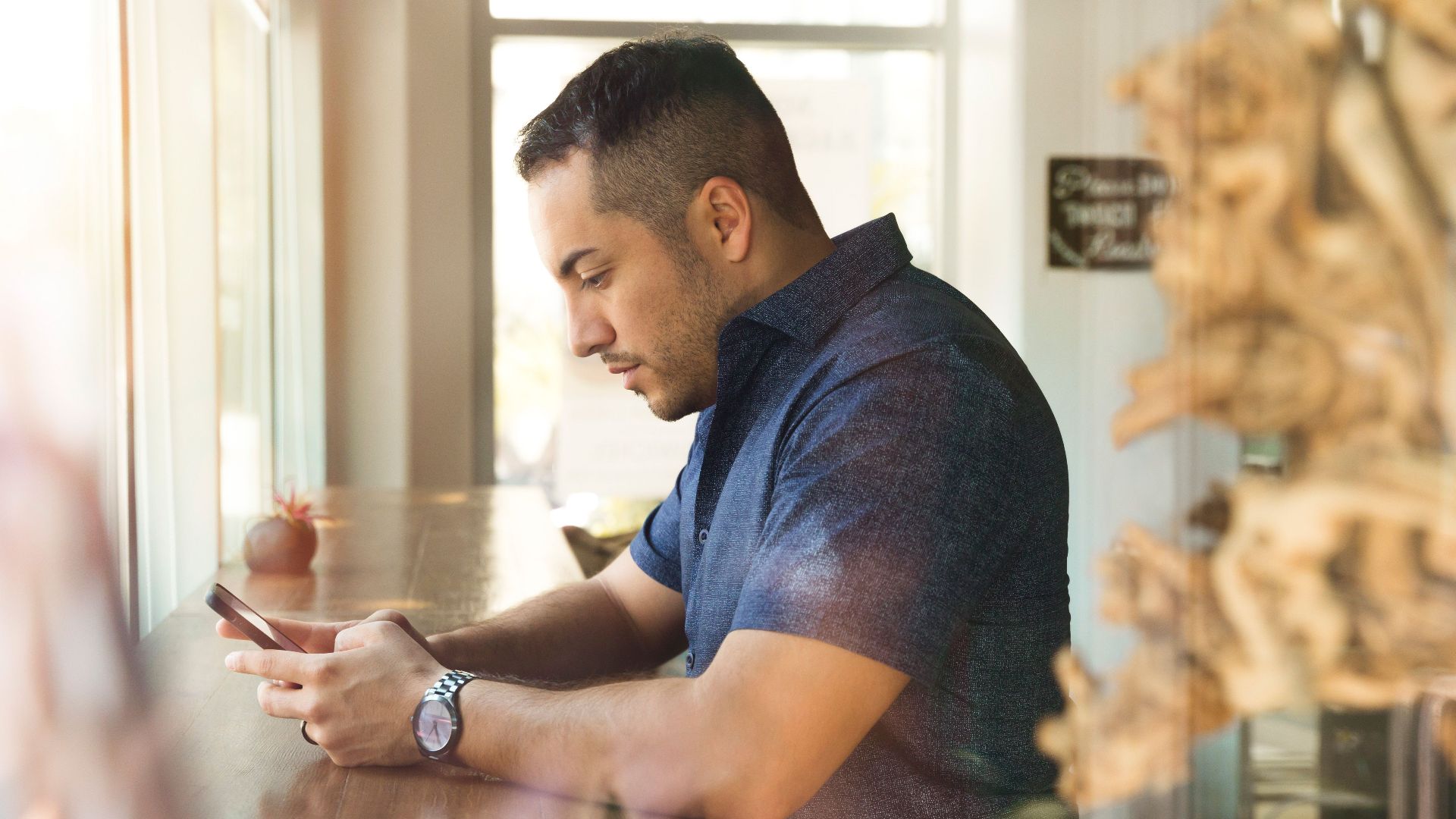 Man in a blue shirt using smartphone indoors by a window at a café.