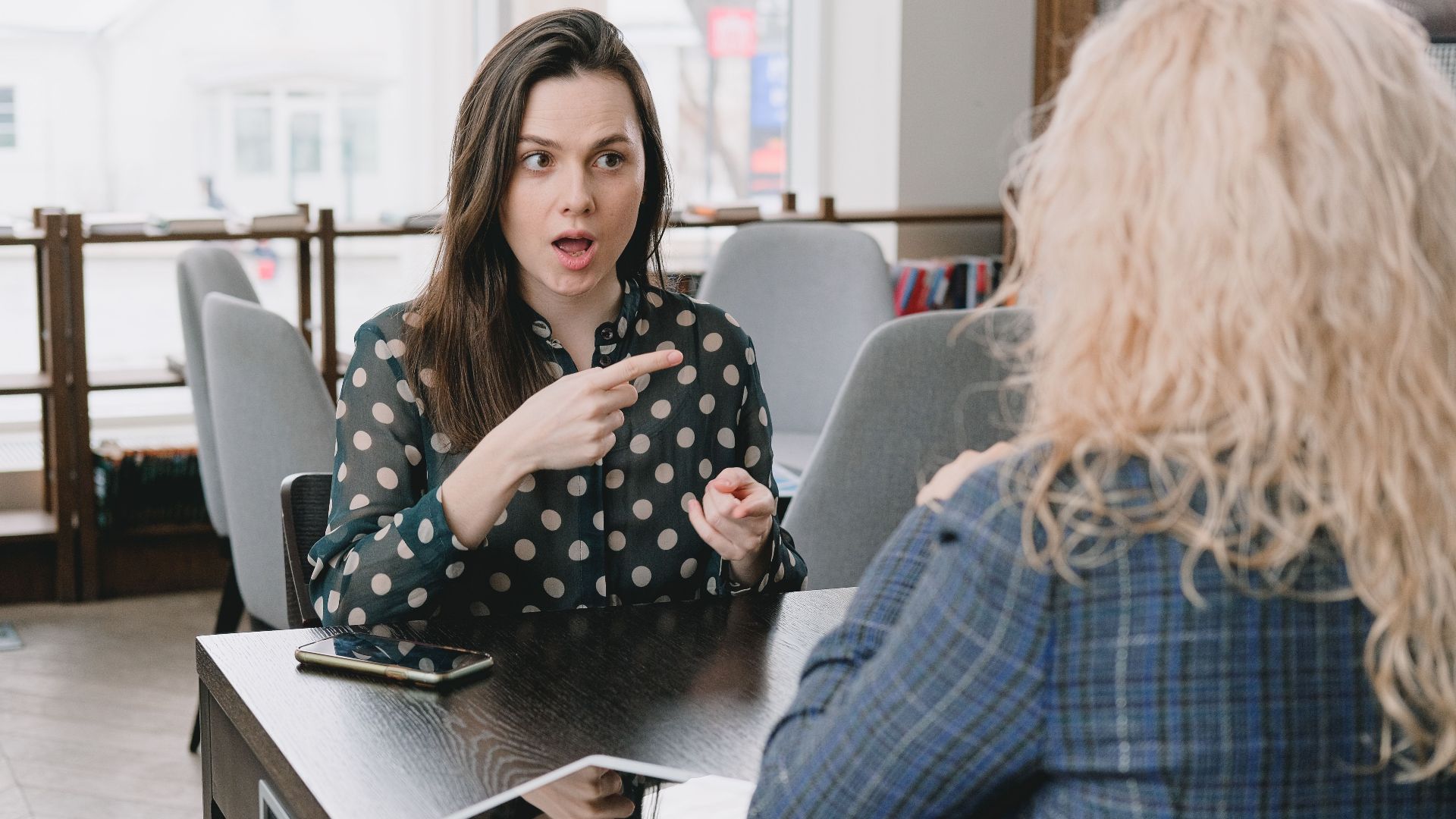 Astonished female pointing away while sitting at table with smartphone and tablet and talking to woman