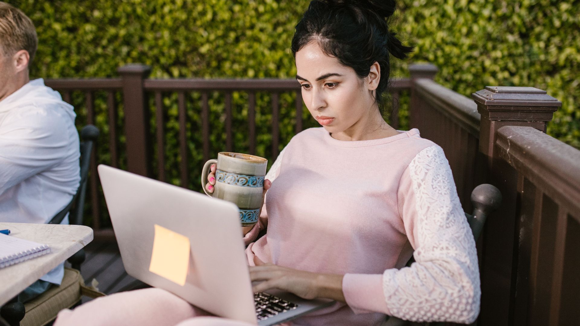 Woman focused on laptop work outdoors, holding a coffee mug. Enjoying remote work in cozy setting.