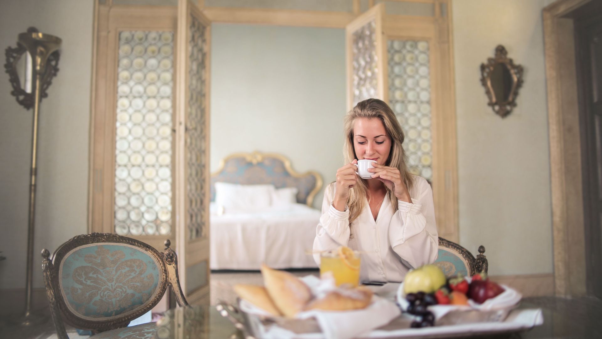 Glad female in elegant wear sitting at table with metal tray of healthy breakfast in vintage hotel room and drinking coffee in morning