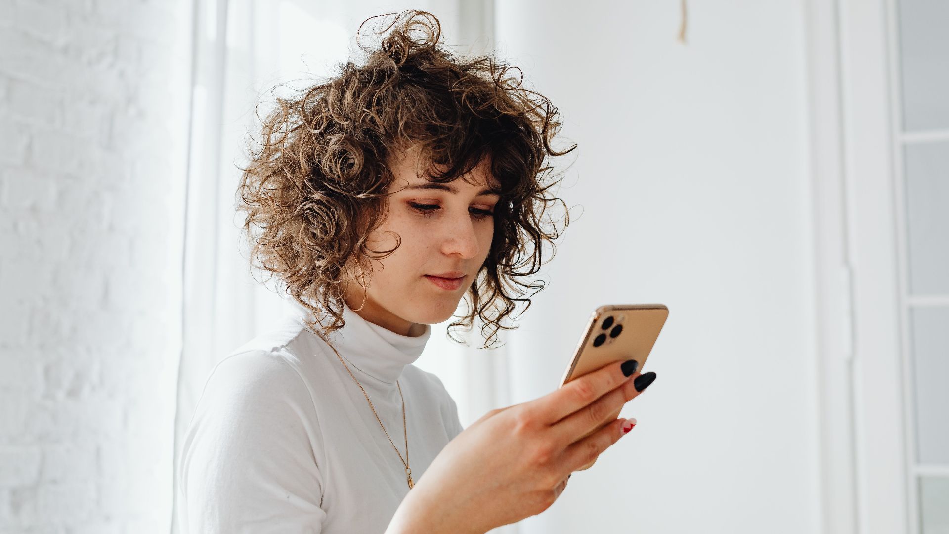 A young woman with curly hair using a smartphone indoors. Natural light and modern decor.
